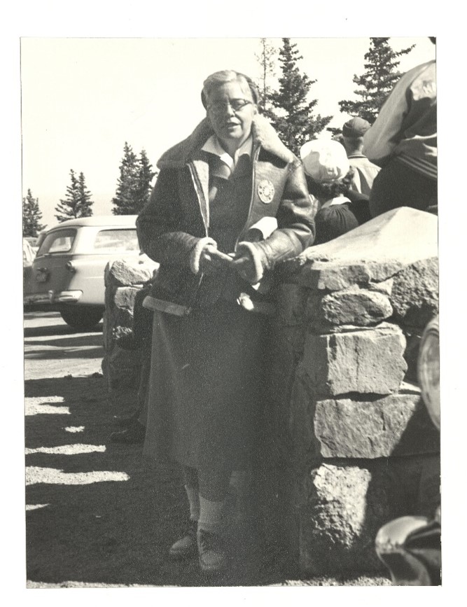 Photo of a woman standing next to a pillar of stacked stones, her left elbow leaning lightly against the top of the stack. A few other people are nearby, facing in the opposite direction. The woman is wearing a below-knee skirt with a blouse and buttoned short jacket, over which she has a leather bomber-style coat. She is wearing a hat on the back of her head, under which her hair is tucked. She is smiling slightly, looking at the person taking the photo.