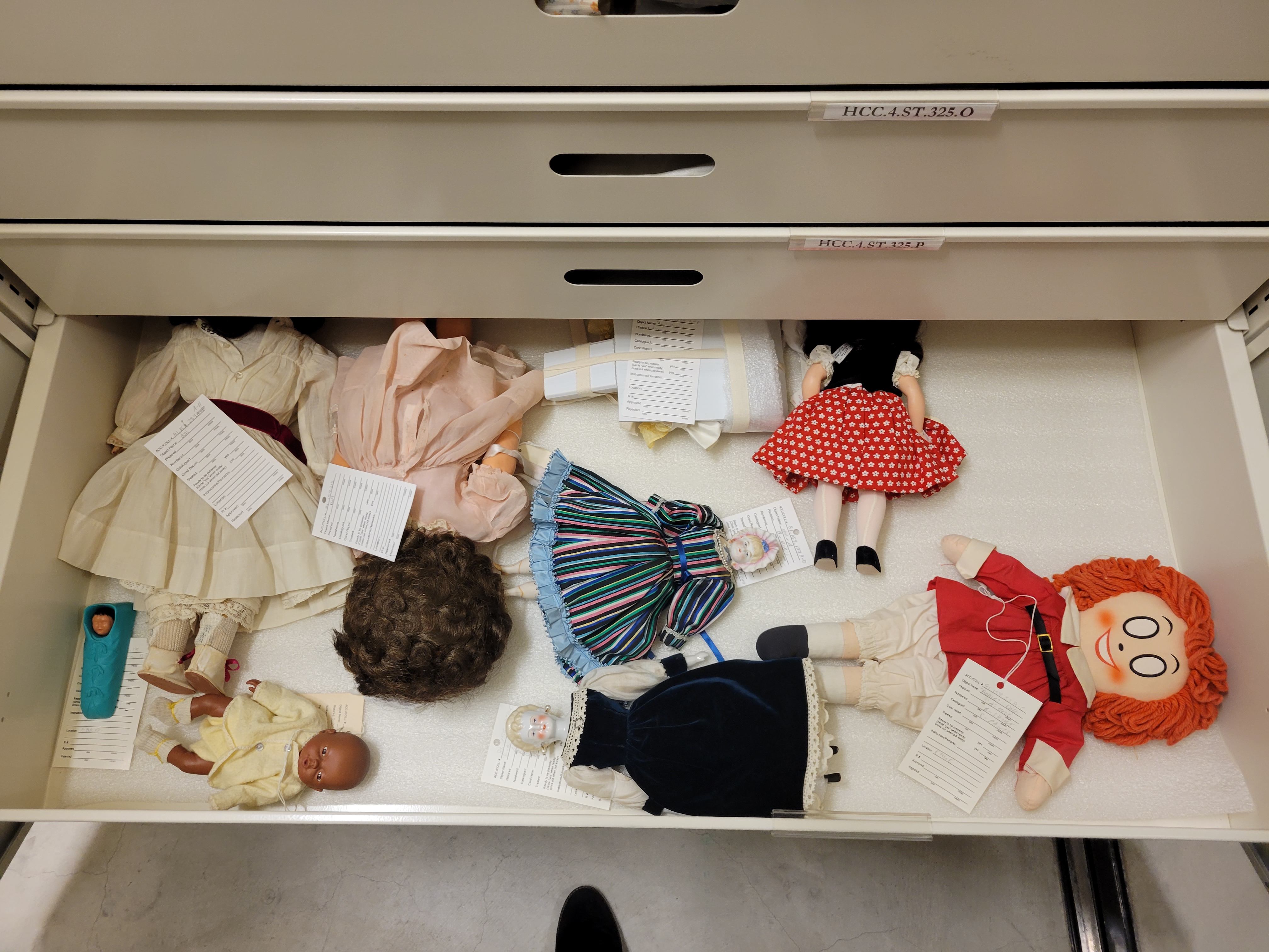 Photo of white metal storage drawers, and the fourth one has been pulled out. Inside the viewer can see several dolls of various sizes, ages, and ethnicities. On the left is a small dark-skinned baby doll, and on the right is a red-haired doll with large white empty ovals for eyes, its face printed on the cloth head. Other dolls in the drawer have fancy dresses in stripes, velvet, and polka dots.