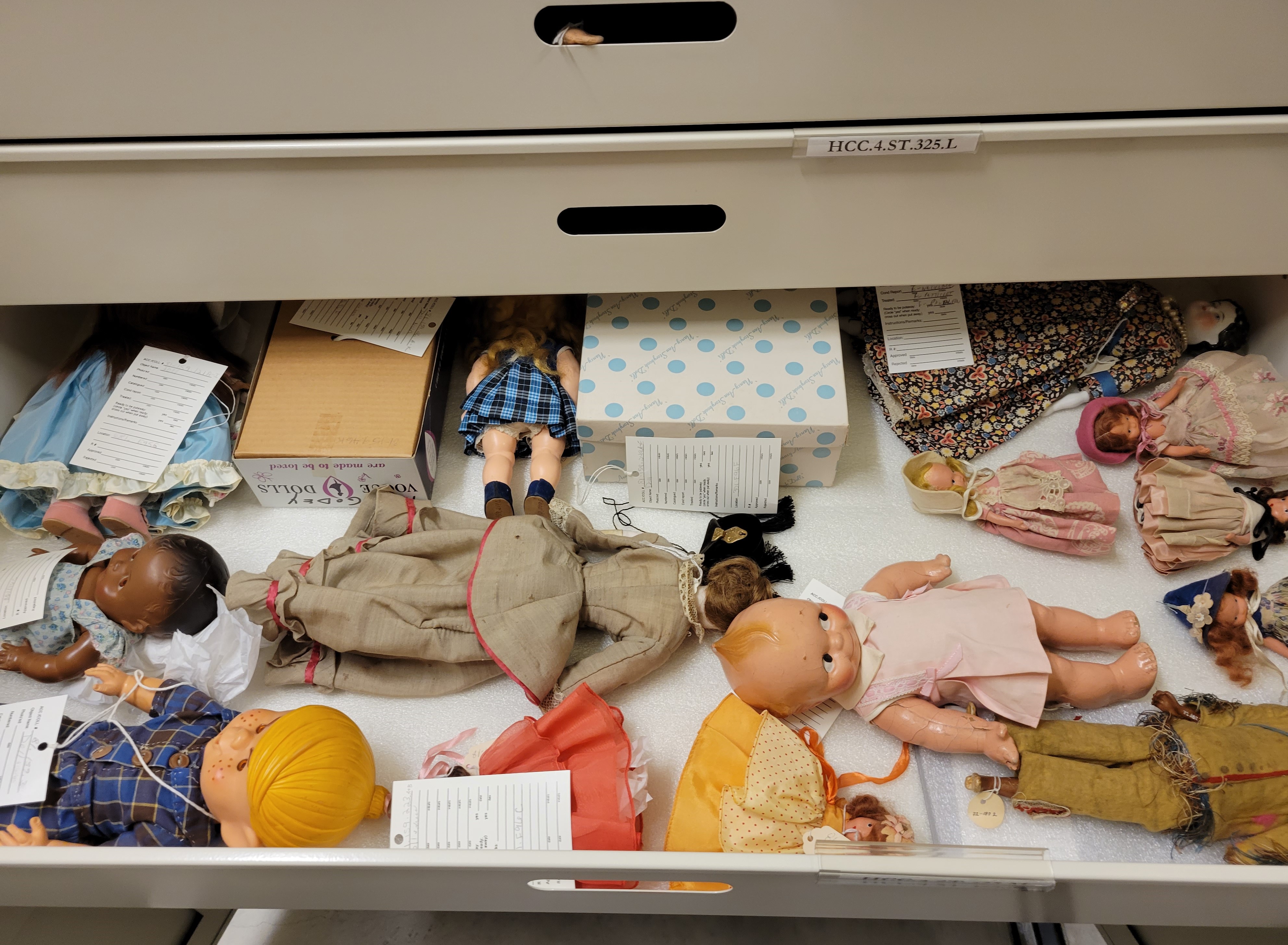 Photo of a white metal storage cabinet with drawers. The third drawer down has been pulled out and it is full of small dolls of different ages, sizes, and ethnicities. In the lower left corner is a plastic Dennis the Menace doll, facing upwards, and on the right amongst the other dolls a Kewpie doll in a pink dress is visible.