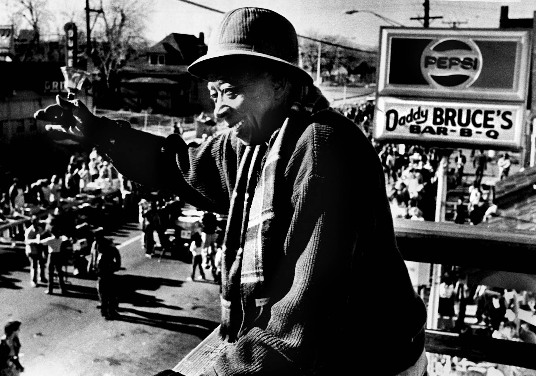 A black and white photograph of Daddy Bruce waving to a crowd. He wears a coat and a hat, and behind him is a sign which reads "Daddy Bruce's Barbecue."