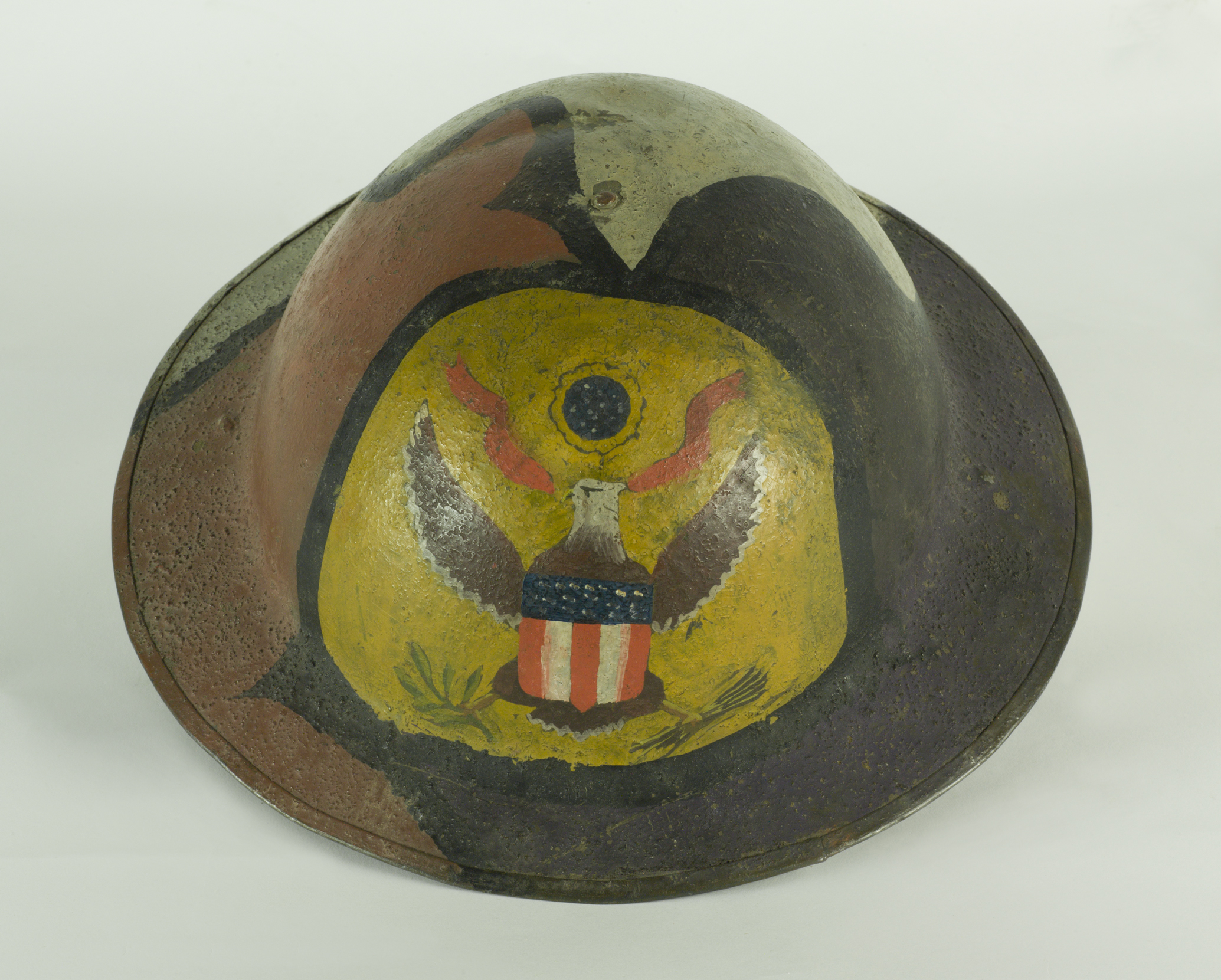 Photo of a Doughboy helmet from World War One. It is a round metal helmet painted with green, brown and black camouflage pattern on the outer surface. A heraldic eagle painted on a yellow background is hand painted on the forehead.