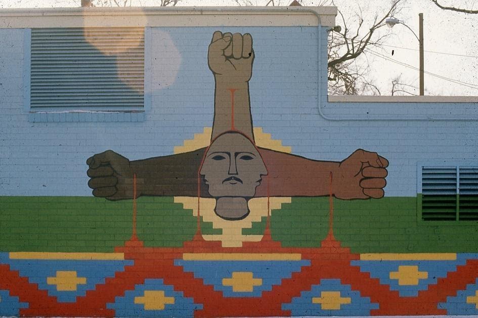 A mural depicts a three-faced head, with three arms of different shades of color coming out of it. Each arm bleeds red, with the red forming part of a geometric design below.