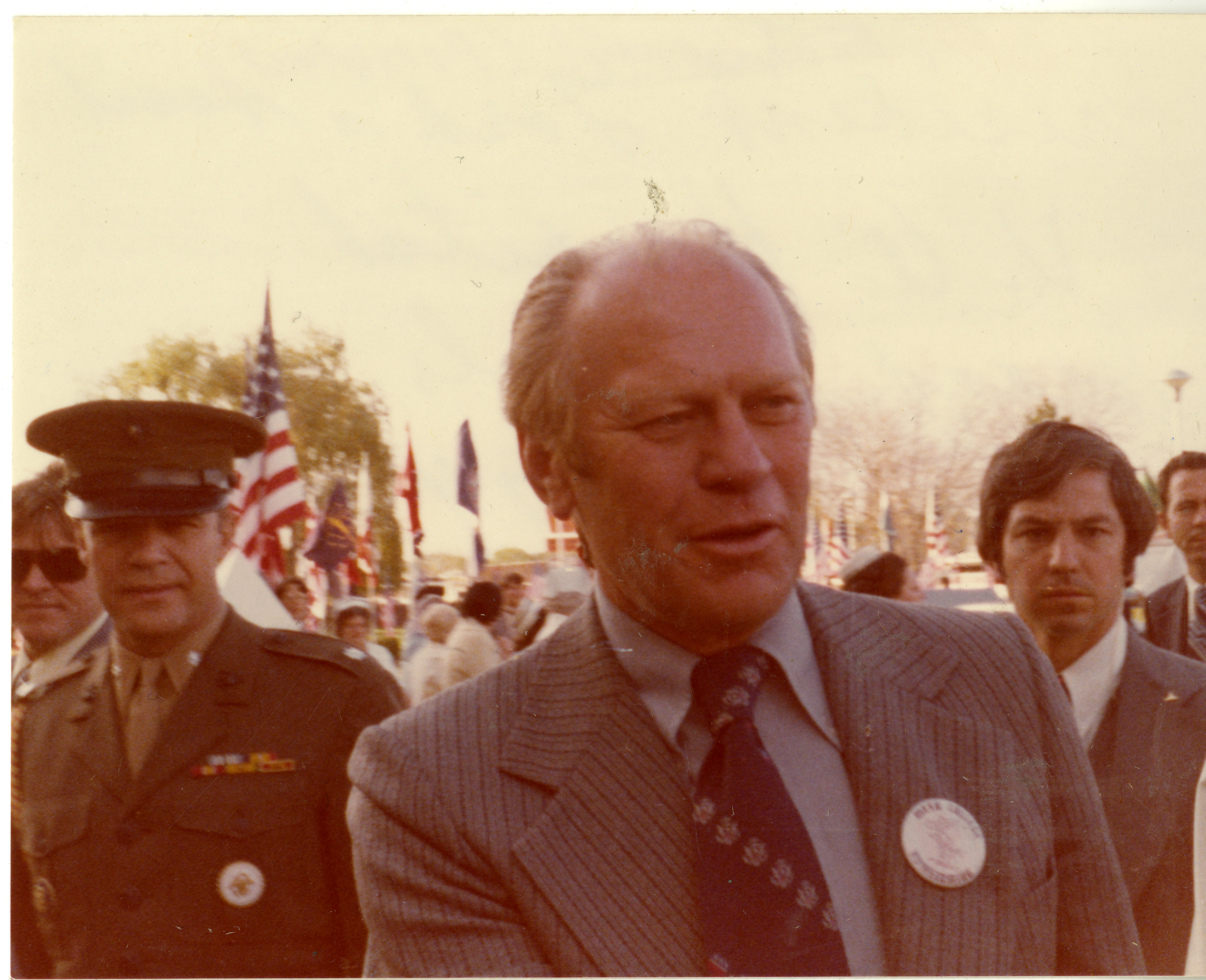 Gerald Ford, wearing a suit and a button, stands with military personnel.