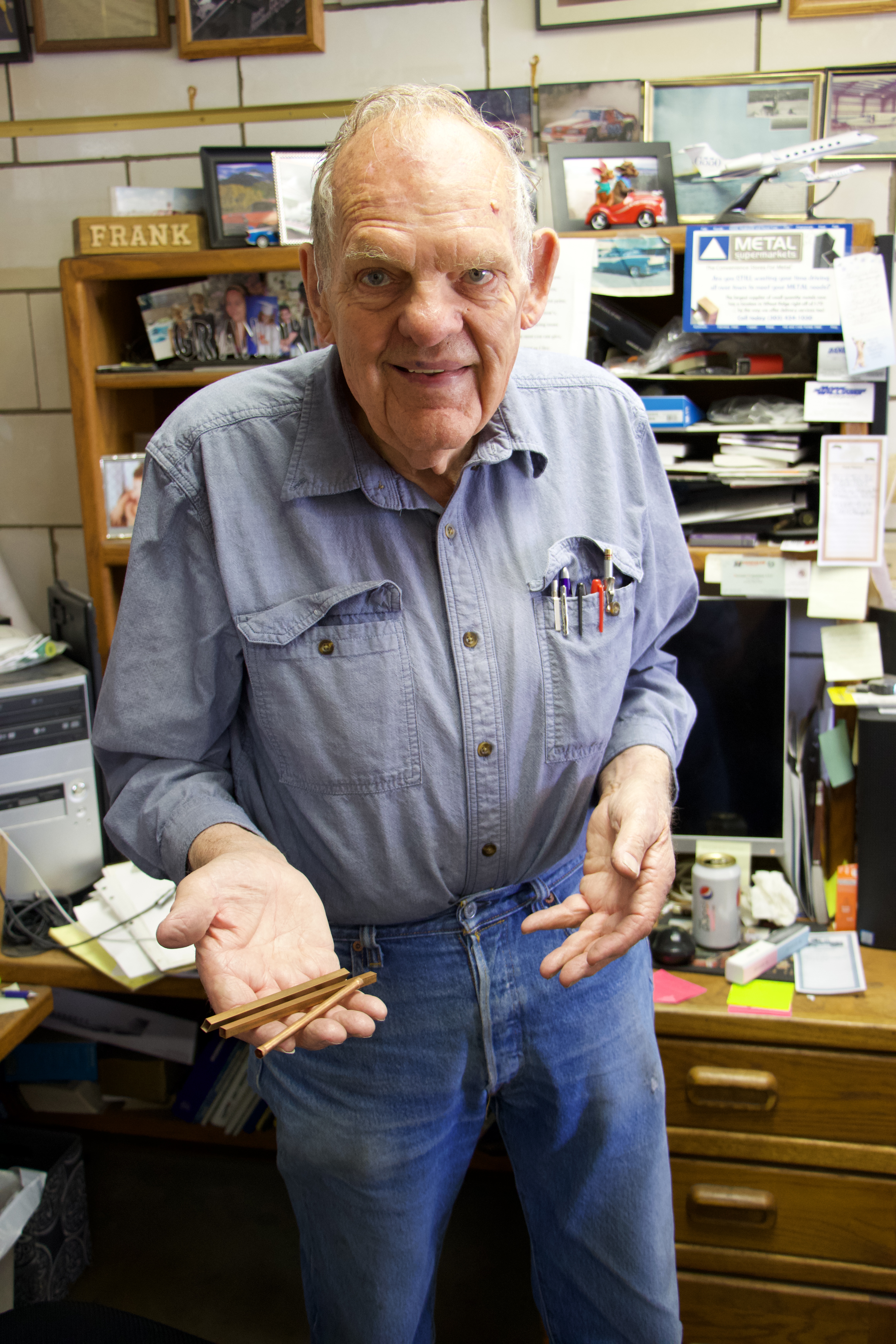 Photo of an older man, standing in front of a desk and shelves. The shelves are full of photos and mementos, and the desk has a computer on it. The man, dressed in blue jeans and a blue denim button-up shirt, with several pens and pencils resting in the breast pocket. He is smiling and holding out small three copper tubes his right hand. The tubes are about a quarter-inch in diameter and about 4 inches long.