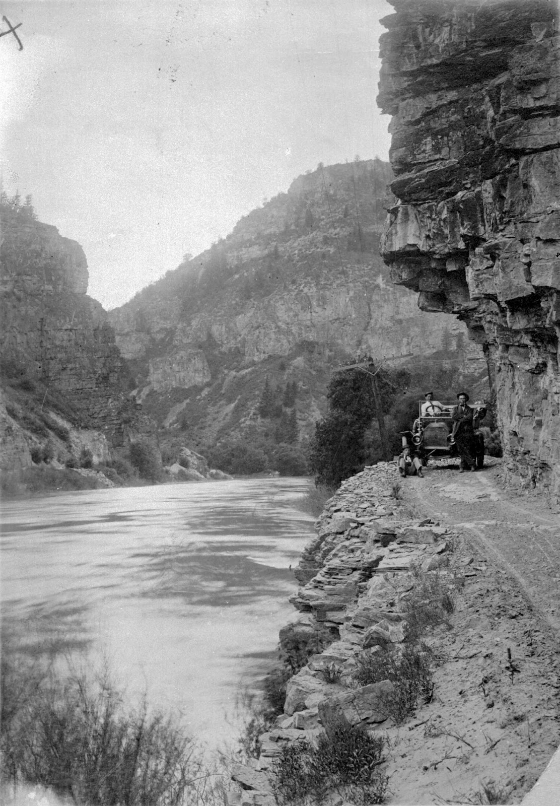 An early 20th-century car drives along a narrow road on the wall of a canyon. To one side of the car is a steep cliff, to the other side is a downward slope ending in water. Behind is a picturesque mountain.