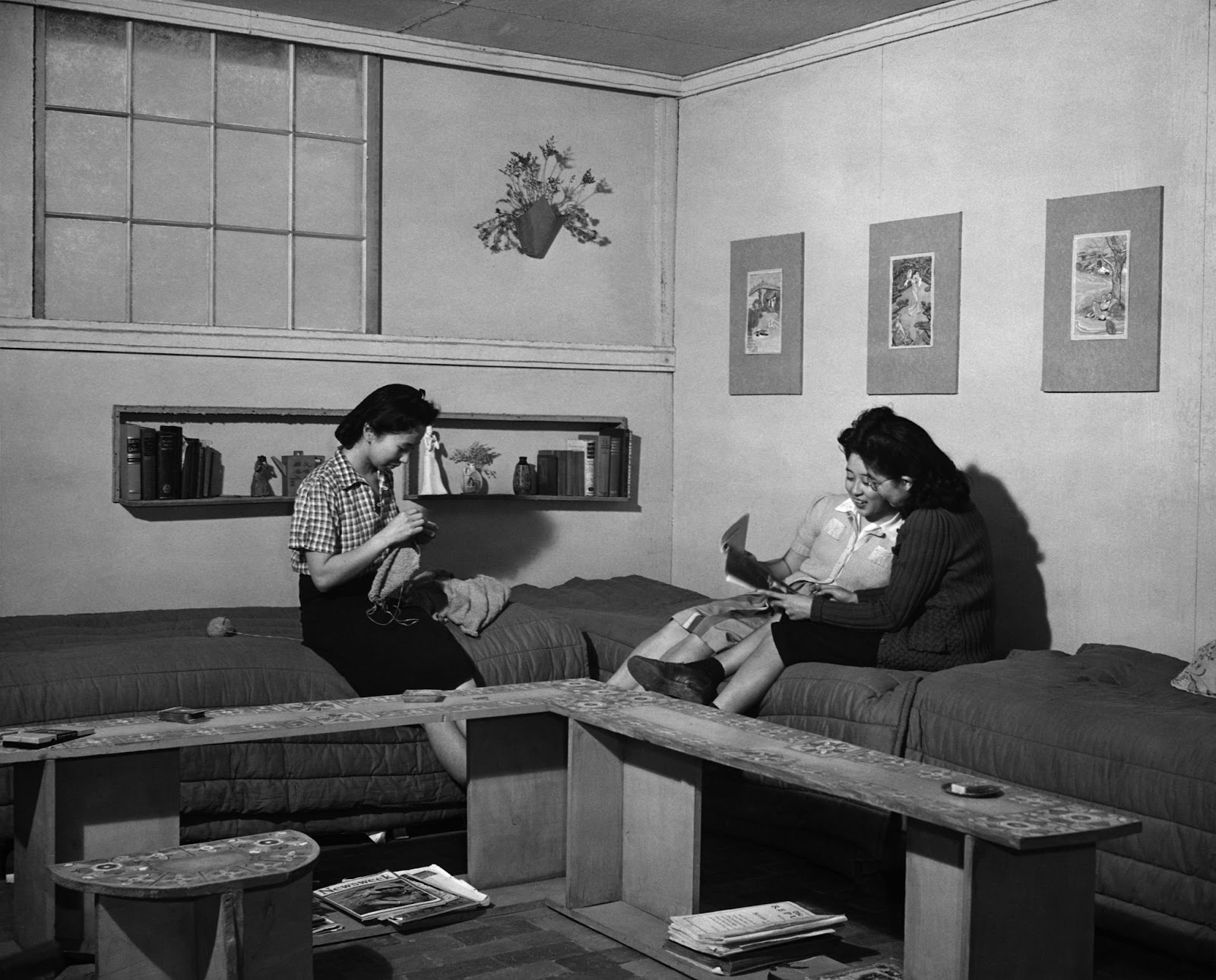 Three women city on beds in a small room. Two are reading a magazine while a third is sewing. Behind them, Japanese artwork hangs on a wall.