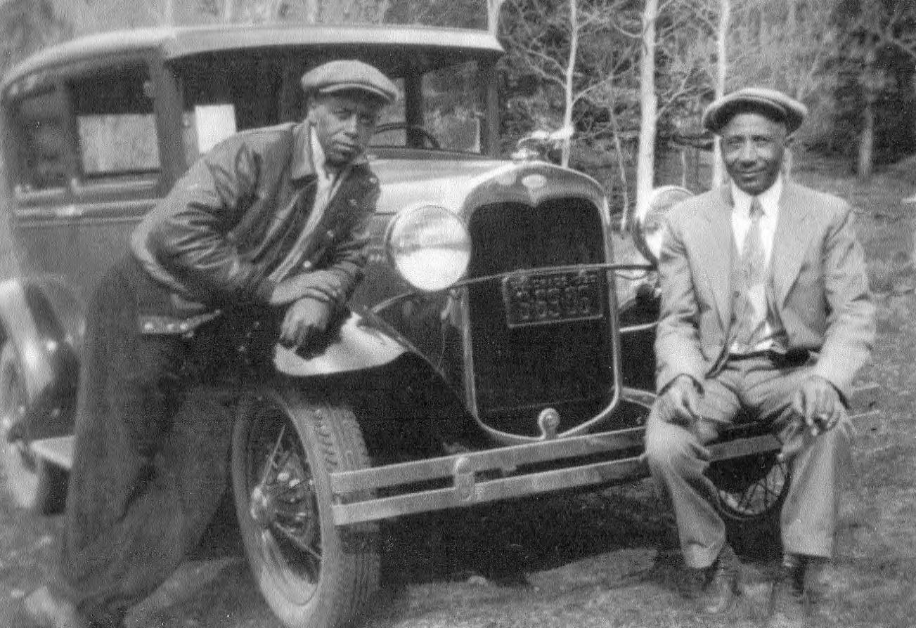 Two African American men stand in front of an automobile, posing for the camera. They wear suits and flatcaps.