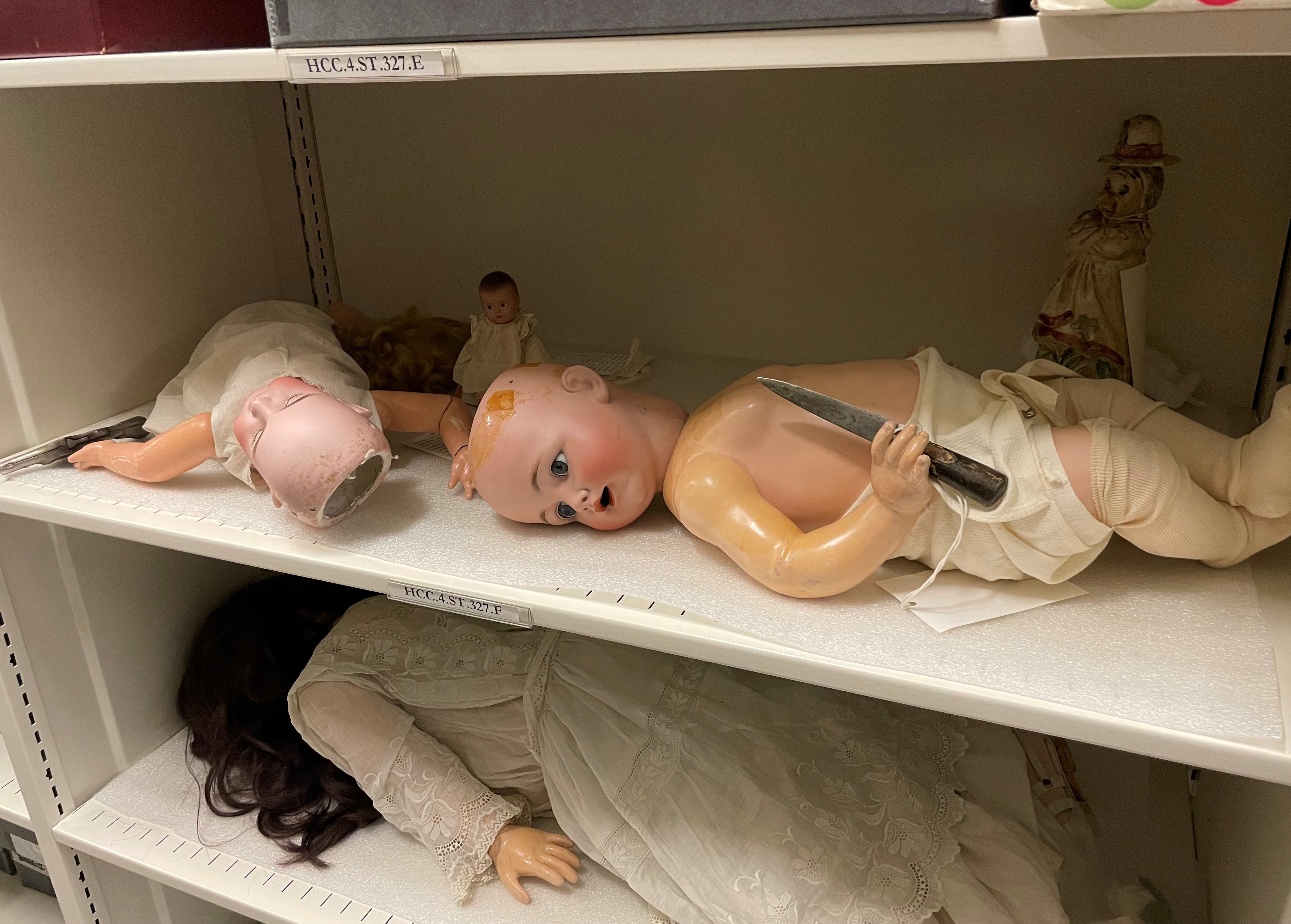 Photo of white metal storage shelves, upon which several dolls are laying either on their backs or facedown. On the higher shelf are two larger dolls that are missing the tops of their porcelain heads and have no hair. The doll on the right, wearing stockings and a cloth diaper, is holding a short knife in its right hand. The knife was staged there as a joke by one of the History Colorado staff members.