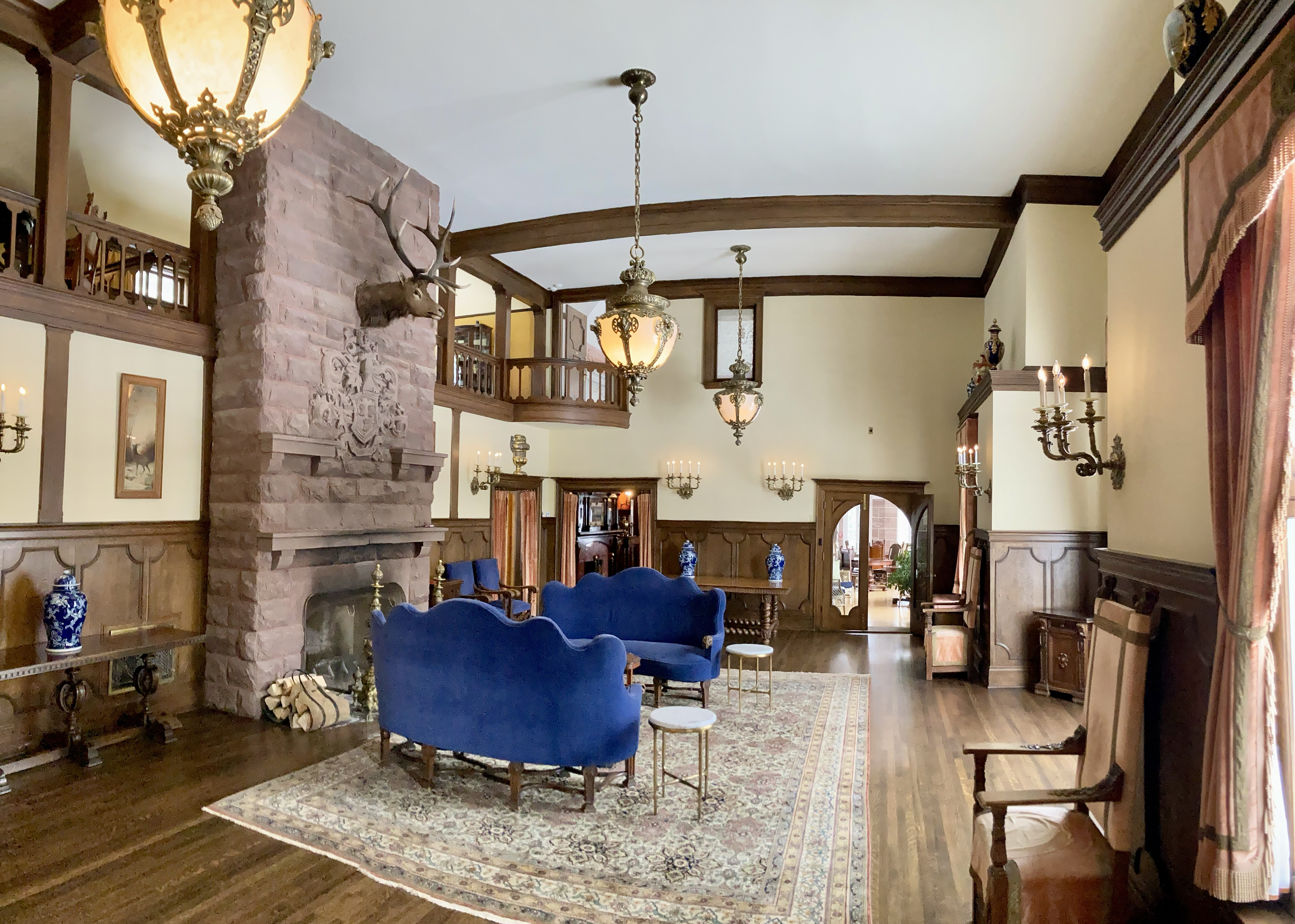 An opulent sitting room with wooden floors, rug, colorful turn-of-the-century furniture, and a stone fireplace.