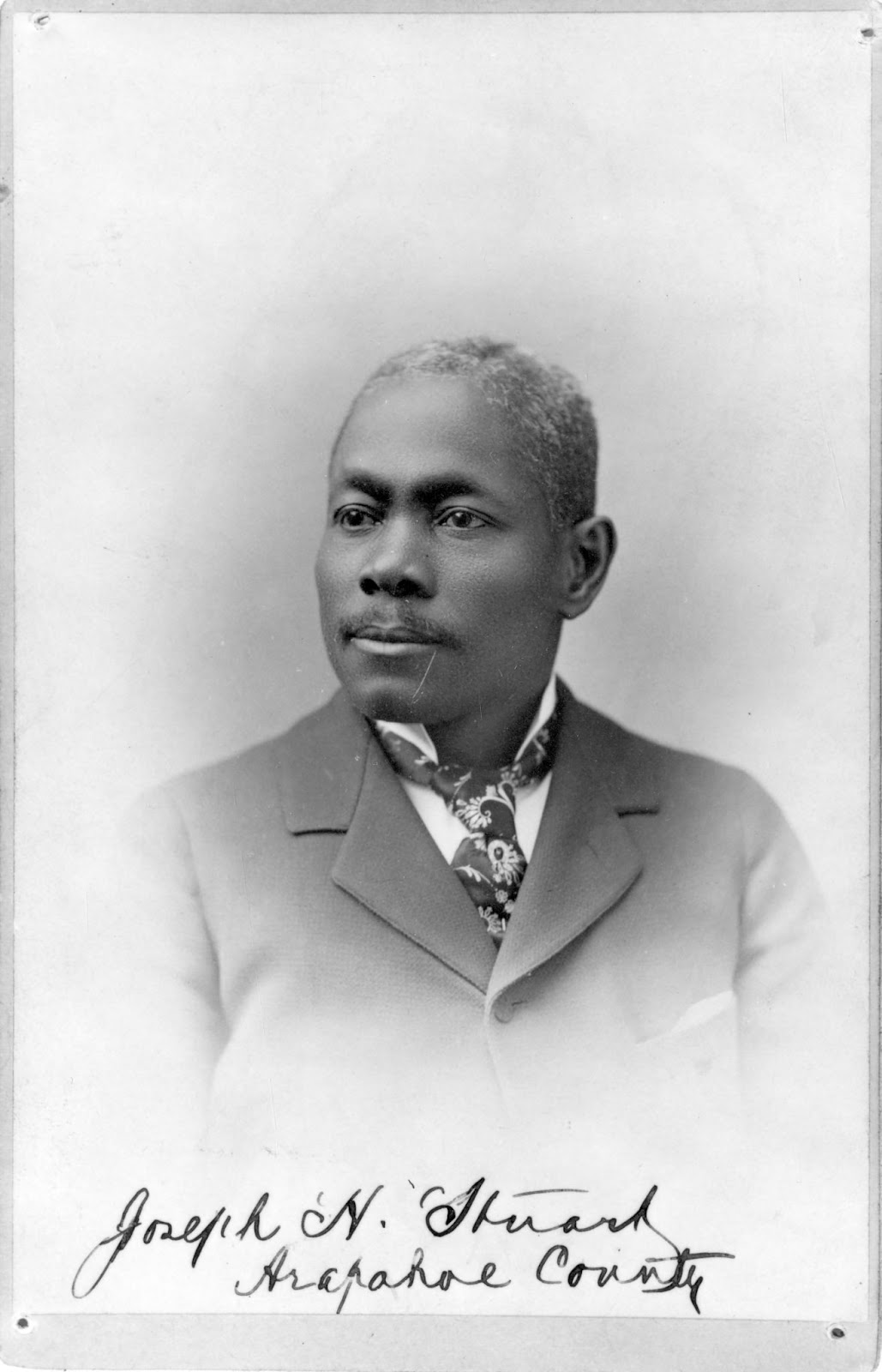 A portrait of a Black man from the shoulders up. He is wearing a 19th century suit. Beneath him is written "Joseph H. Stuart, Arapahoe County".