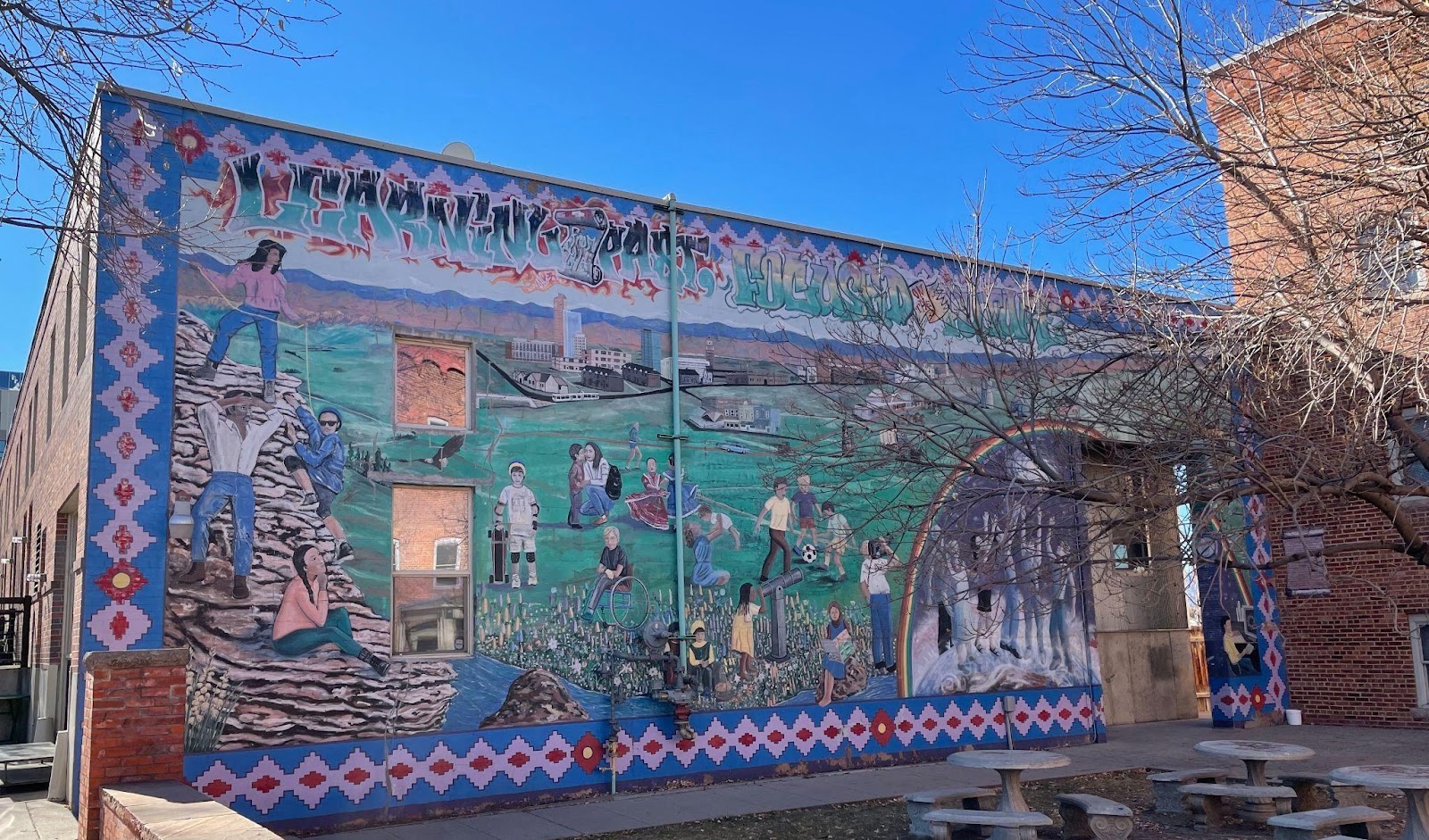 An image of the mural Learning from the Past, Focused on the Future, which depicts many Chicano individuals doing many different activities. The mural is painted on the side of a business building.