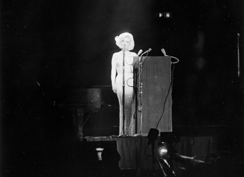 Photo in black and white of Marilyn Monroe, standing on stage next to a podium, singing. Her figure is illuminated against the darkened stage by a single spotlight, highlighting the iconic "nude" and rhinestone encrusted dress that she wore for the occasion.