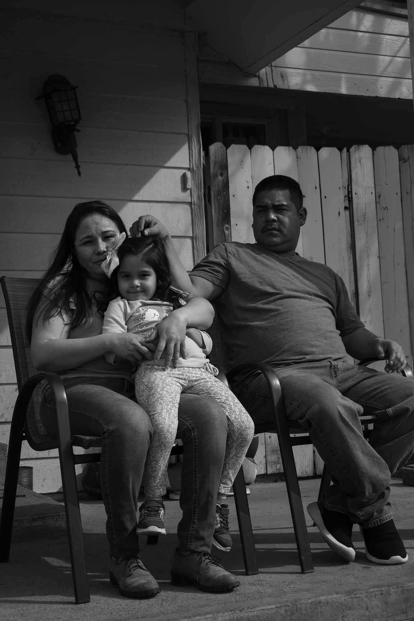 A family sits on a porch. A man sits on a chair to the right, while a woman sits on a chair to the left with a young girl in her lap.