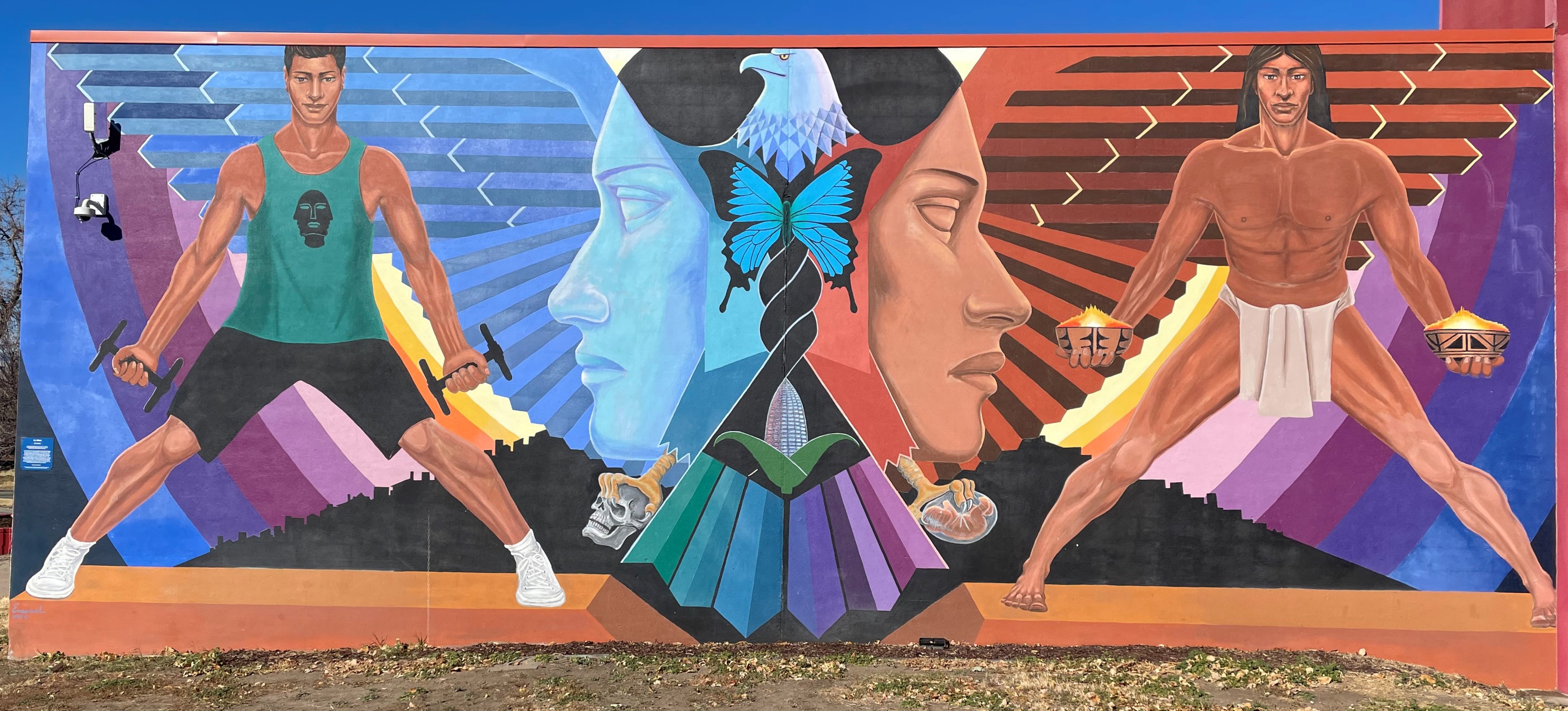 A colorful mural depicting an eagle whose wings form the faces of Chicana women. On one side is a modern chicano athlete, on the other side is an Indigenous person participating in a ritual.