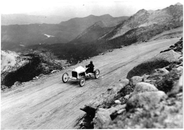 Photo of an open-wheeled race car, driving up the dirt track of Pikes Peak. The rocky hillside is in the foreground, and in the distance, tall mountains loom. The track itself is only lined with a few naturally occuring small rocks, and otherwise drops off down the hillside. The car in this historic photo has a small engine housing, and otherwise the driver sits on an open seat, dressed in a coat and helmet and exposed to the challenges of the race track. 