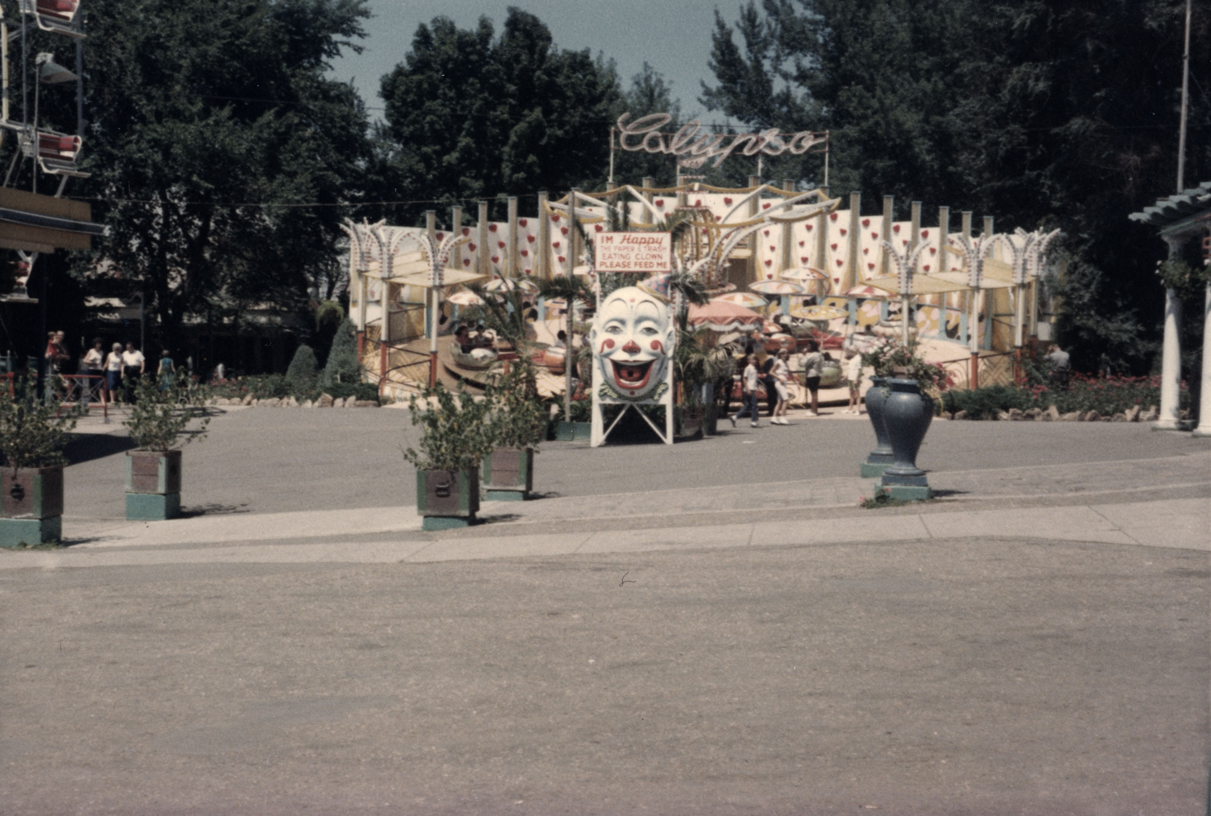 Photo of Elitch Gardens. In the background is the Calypso ride, which is stationary in this image and a few people are walking toward the gate to enter the ride. In front of the ride is a large white clown face, with its mouth open wide and its large red tongue has a large hole in the center. The clown is staring straight ahead. Above the face is a sign that reads "I'm Happy the Paper & Trash Eating Clown. Please feed me."