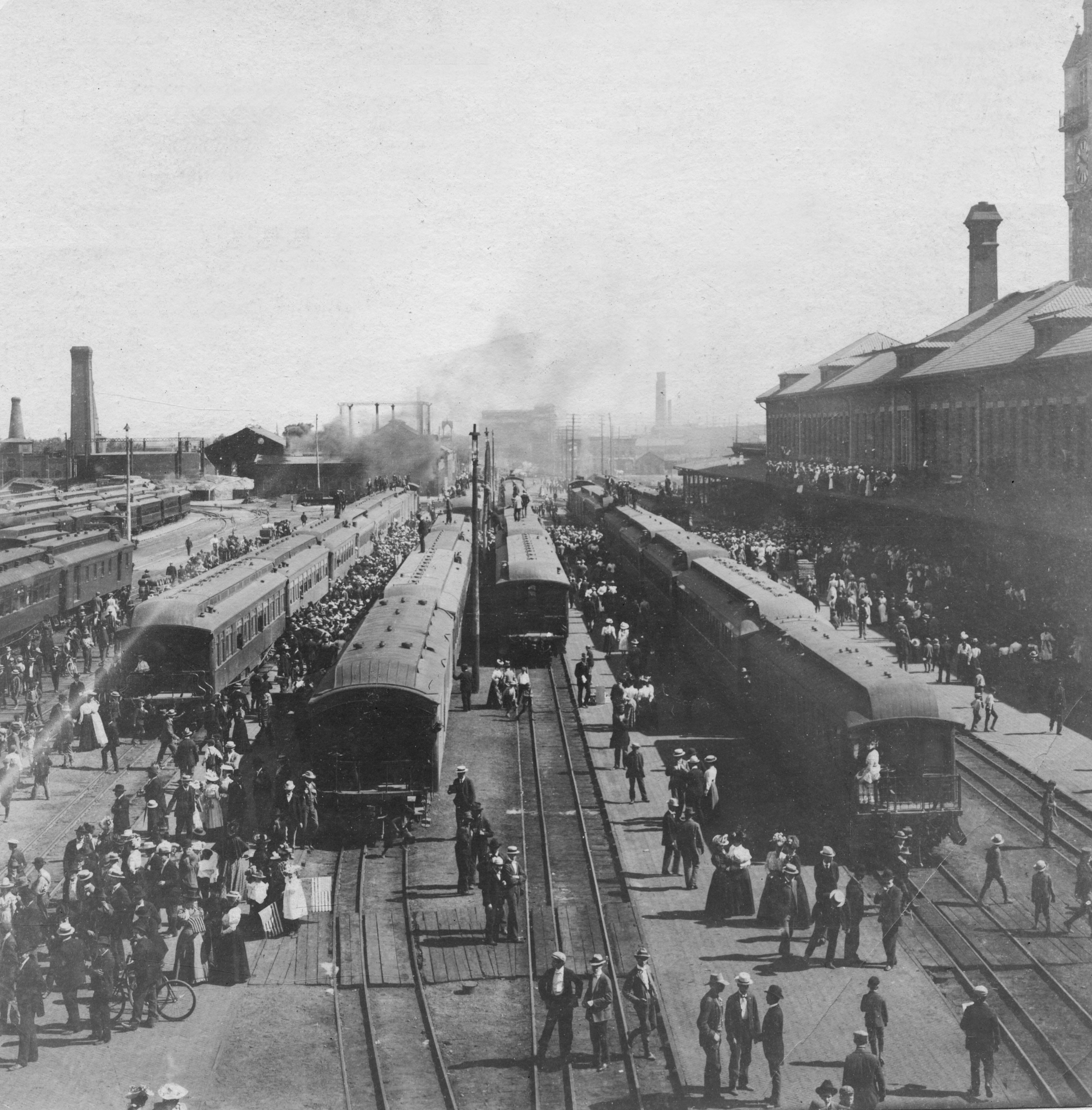 Photo of several trains stopped along several lines of train tracks. Crowds of people, most wearing brimmed hats and either suits or skirts, are gathered around the trains to board. A few people are pushing bicycles. At the end of the tracks, black puffs of smoke can be seen rising in the air. The long train depot building is on the right of the photo.