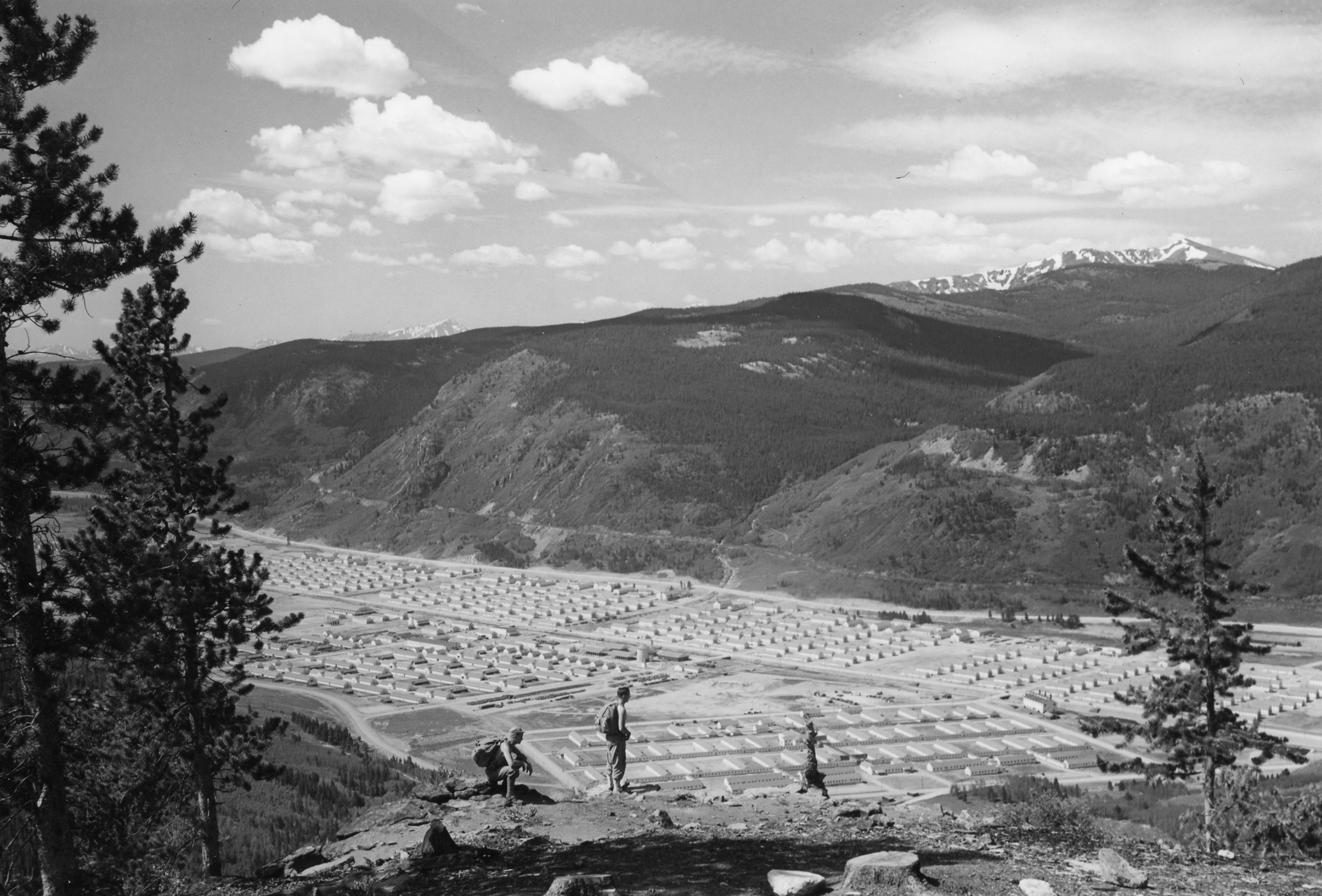 A view of Camp Hale from a nearby mountain, the entire camp visible. A group of men are looking down at the camp from a rise.