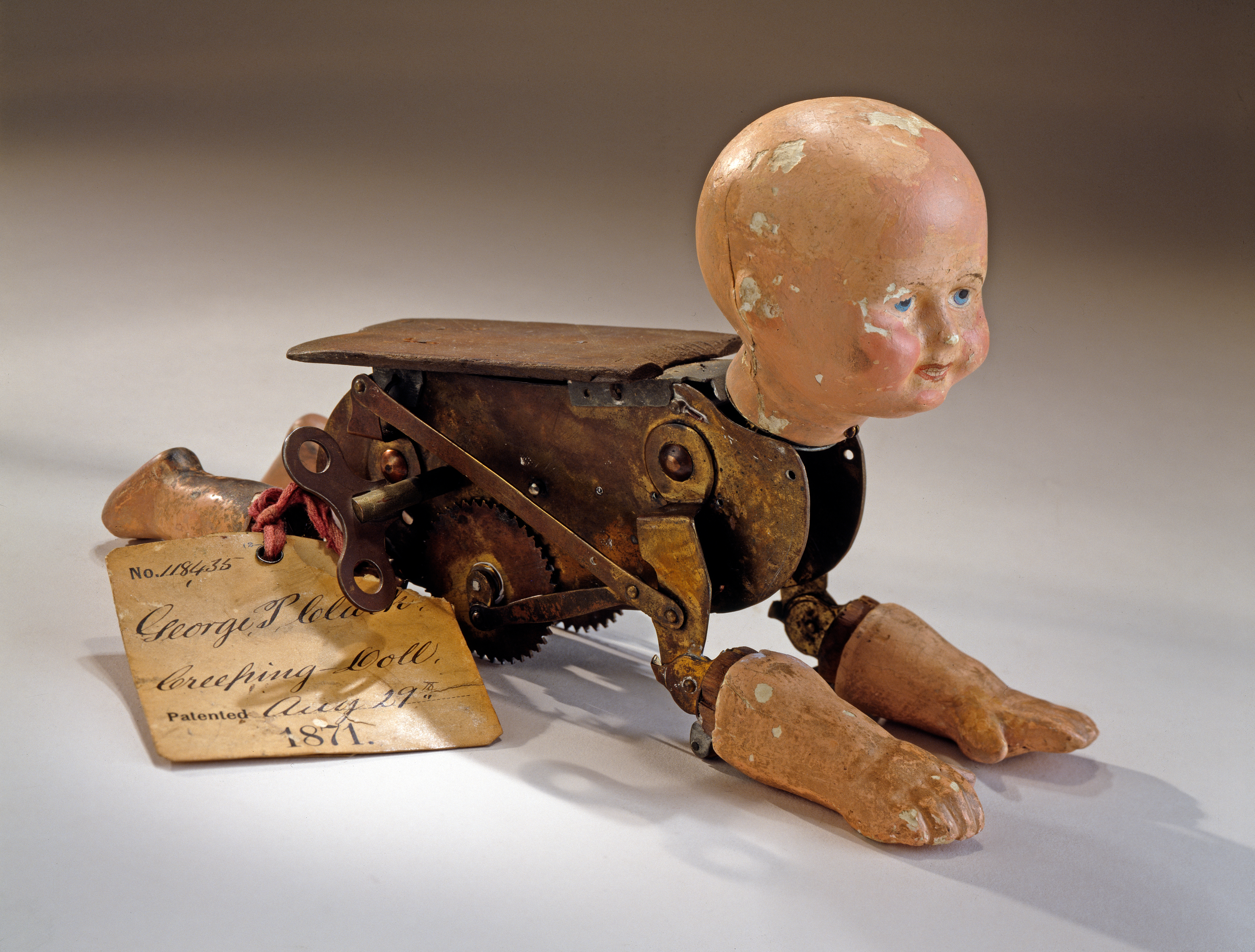 Photo of a mechanized doll which appears poised to crawl across the white tabletop. The doll has no hair, rosy cheeks, and blue painted eyes. Its head is attached to the metal workings that make up the body of the doll, upon which porcelain hands and feet have been attached. The clockwork gears and key are on the doll's side, along with a tag which has handwriting that says "No. 118435. George P. Clark. Patented Aug 29, 1871."