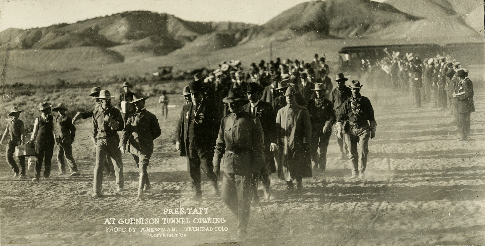 A group of men walk across a dusty landscape wearing coats and hats. President William Howard Taft is one of their number. Mountains are visible behind them.
