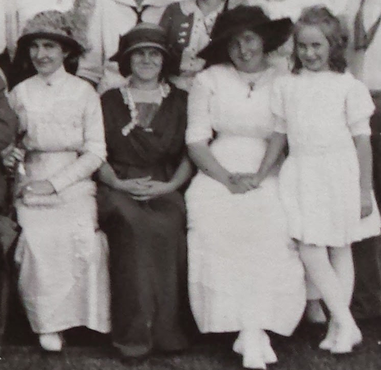 Three women in turn-of-the-century dress sit together in an outdoor setting. The second from the left is wearing a dark dress, while the other three wear light colored or white dresses. The three older women are wearing broad-brimmed hats.