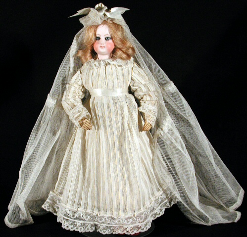 Photo of a doll, poised upright in front of a black background. The doll is female, dressed in a fancy white dress with a long full skirt and long sleeves with a lacy collar and a ribbon waistband. The dolls long red hair is topped with a long tuile veil and a bow, and her porcelain face is full with rosy cheeks and eye makeup.