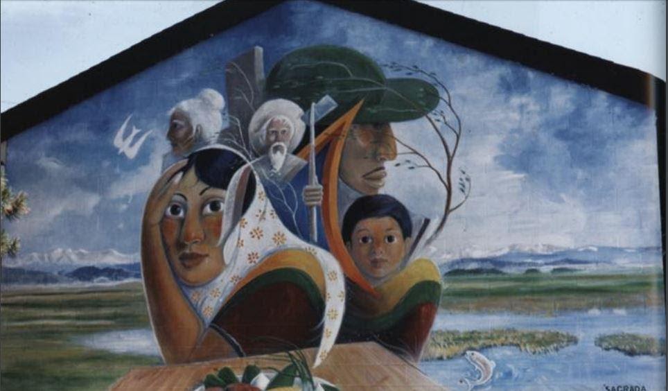 A mural depicts the any faces of a chicano family, of all ages and gender. They are surrounded by verdant farmland, and there are mountains in the distance.