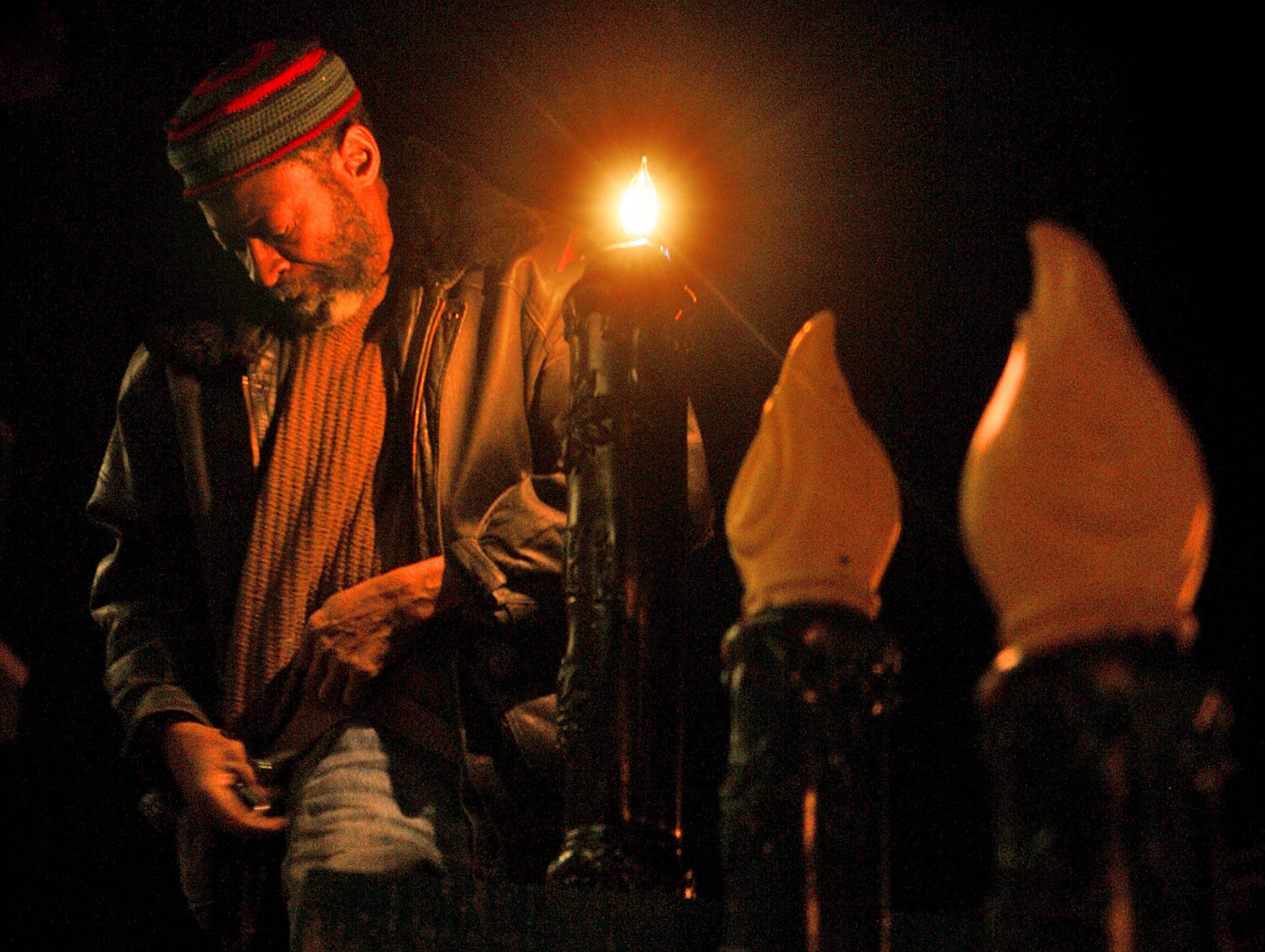 Photo of an African American man standing next to tall candle ornaments , the body of which have been painted black and the reproduction "flames" are yellow. The tallest candle decoration has a bare light bulb where the "flame" cover should go, and the man is testing the light bulbs to make sure they are functioning properly. He is wearing a crocheted cap in with red, black, and gray horizontal stripes, and a black leather bomber-style jacket with jeans and a golden yellow sweater. It is night time.