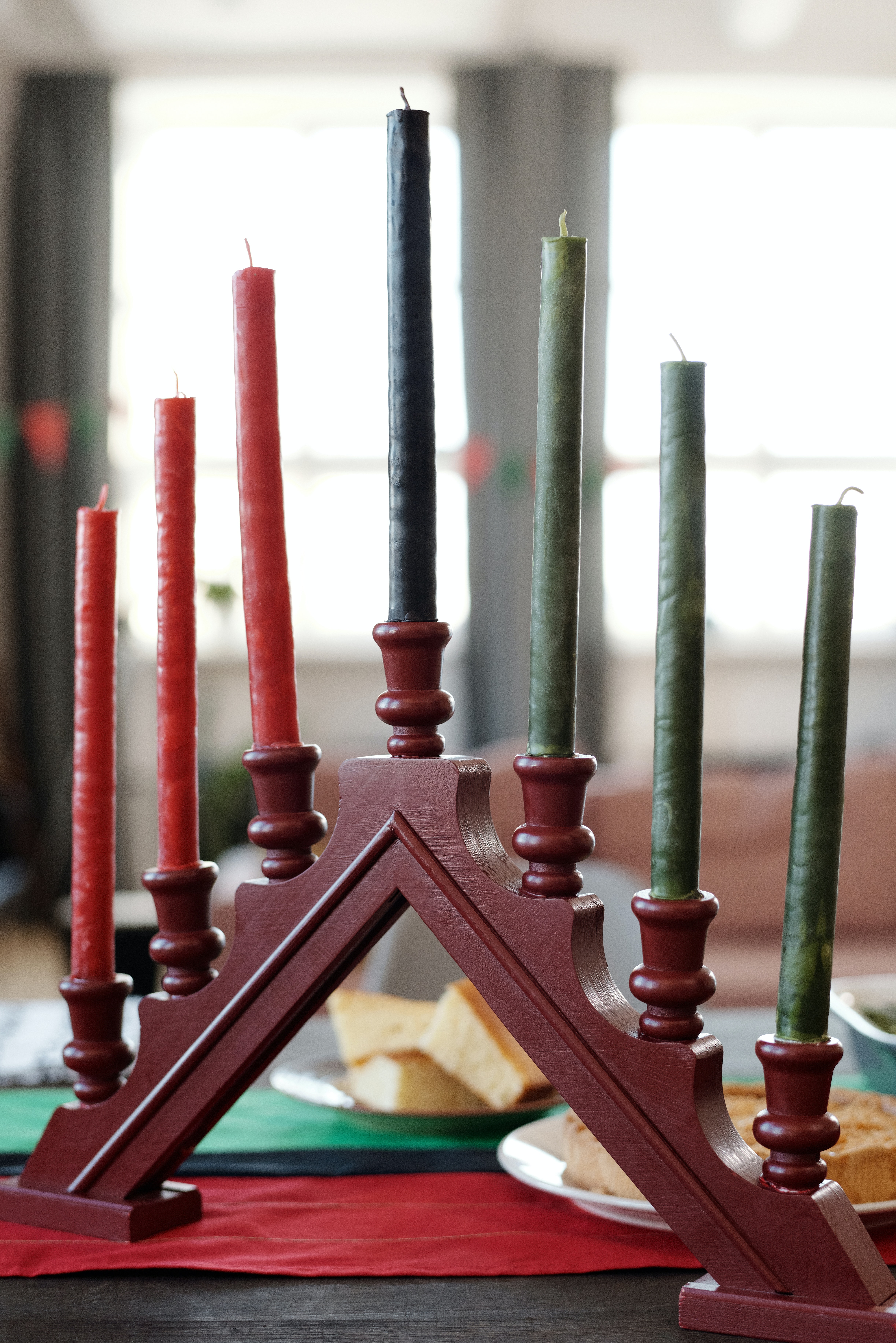 Photo of a wooden candelabra that is painted red. It has holders for seven candles and is shaped in an upward triangle so that the fourth (middle) candle is the highest, with the other candlesticks descending down to the base. The center candle is a black taper, while the three candles on the left are red tapers, and to the right are three green tapers. The holder sits on a table, with plates of food barely recognizable in the background. In the far background are tall windows emitting daylight.