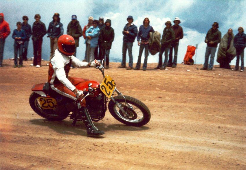 Photo of a motorcycle racer, speeding along a dirt race track while spectators line the edge to the track to watch. The rider is dressed in a red and white race suit with a red helmet covering the whole head, and black leather boots with buckles for closure. The motorbike has two yellow signs denoting its race number, 25, on the front of the handlebars and also in the center of the rear wheel. 