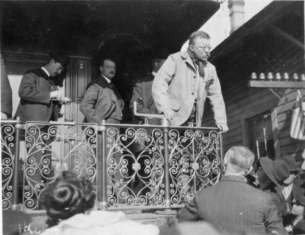Theodore Roosevelt stands at the backplate of a caboose, looking down at people on the train platform below. He is wearing a duster. Behind him stand men in suits.