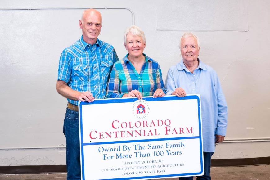 Three older individuals, one man and two women, pose with a large certificate which reads "Colorado Centennial Farm: Owned by the Same Family for More than 100 years."