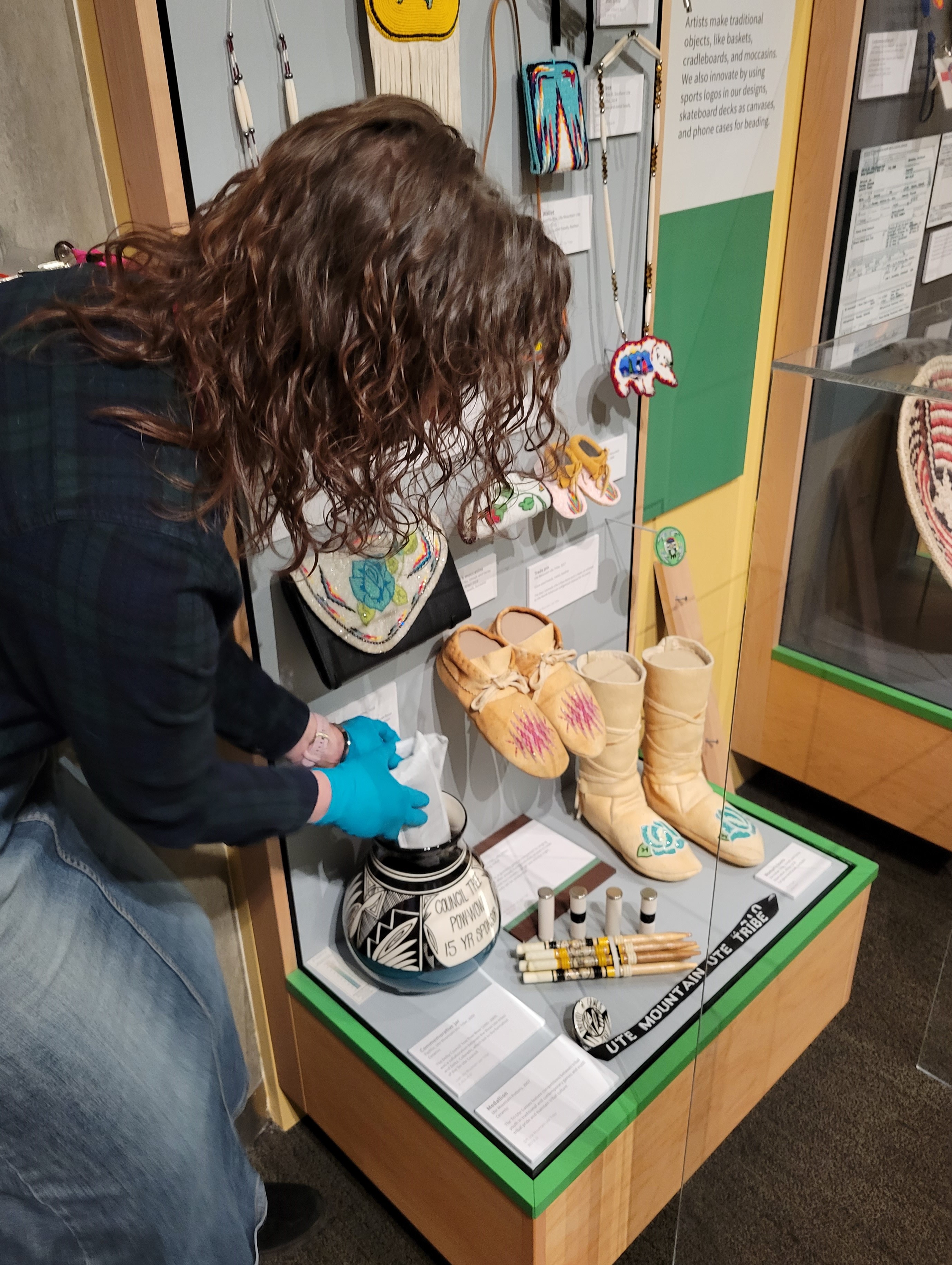 Photo of a person leaning down to change a sachet of activated carbon that has been discreetly placed inside of a pot that is part of an exhibit of Native American artwork from the Ute Mountain Ute Tribe. The museum employee changing the carbon is wearing blue nitril gloves and carefully places the sachet inside of the glazed pot.