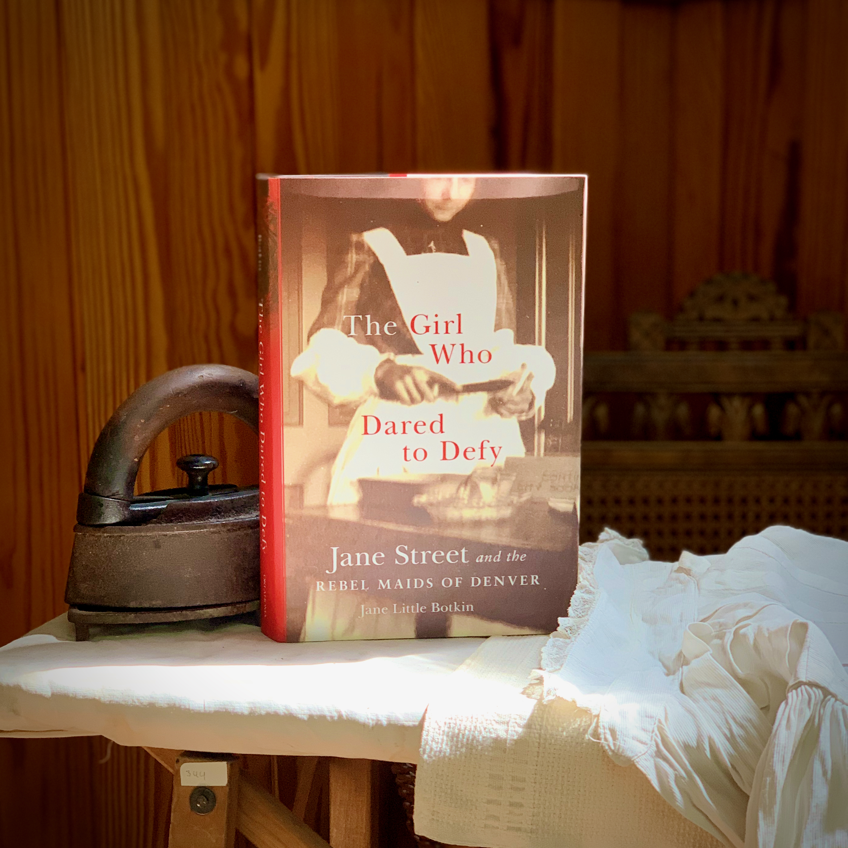 Photo of a book sitting on an ironing board, next to an antique iron and a white garment. Titled "The Girl Who Dared to Defy: Jane Street and the Rebel Maids of Denver," the cover is a black and white image of a female who wears a dark colored dress over which a long white bibbed apron is worn. She is standing in front of a table with a shallow small bowl and what appears to be a rectangular cooking pan. Although only part of her face is seen, she looks down at an open book she holds in her hands.