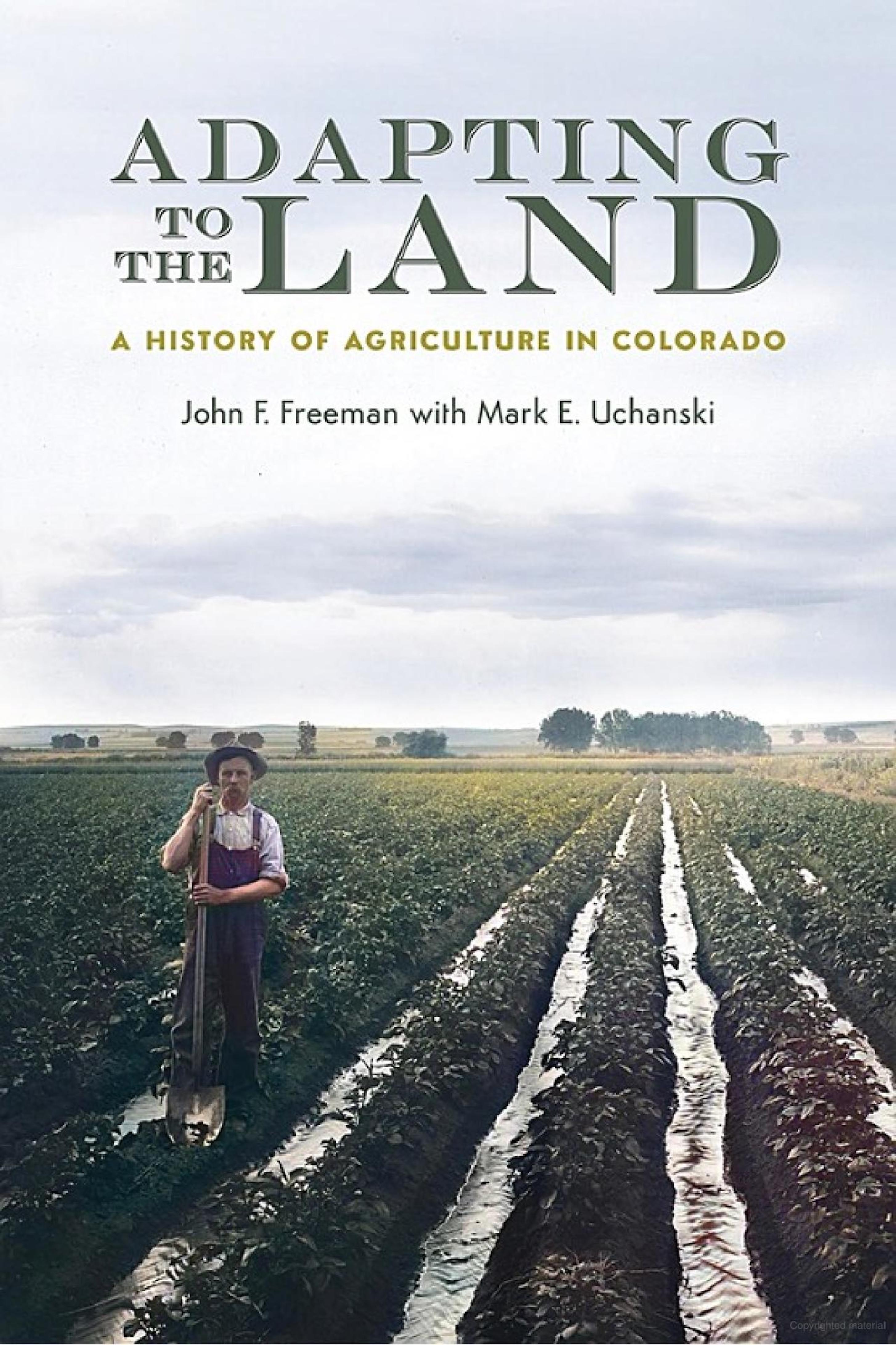 Photo of a book cover whose title is "Adapting to the Land: A History of Agriculture in Colorado." Below the title are the author' names: John F. Freeman with Mark E. Uchanski. The photo on the cover is across the cover's entirety from edge to edge, and is what appears to be an old photo, recolored, of a farmer stanging amongst rows of crops that stretch off far into the distance. Short leafy plants cover the linear mounds of early, and water flows between the mounds. The farmer is standing with a shovel.