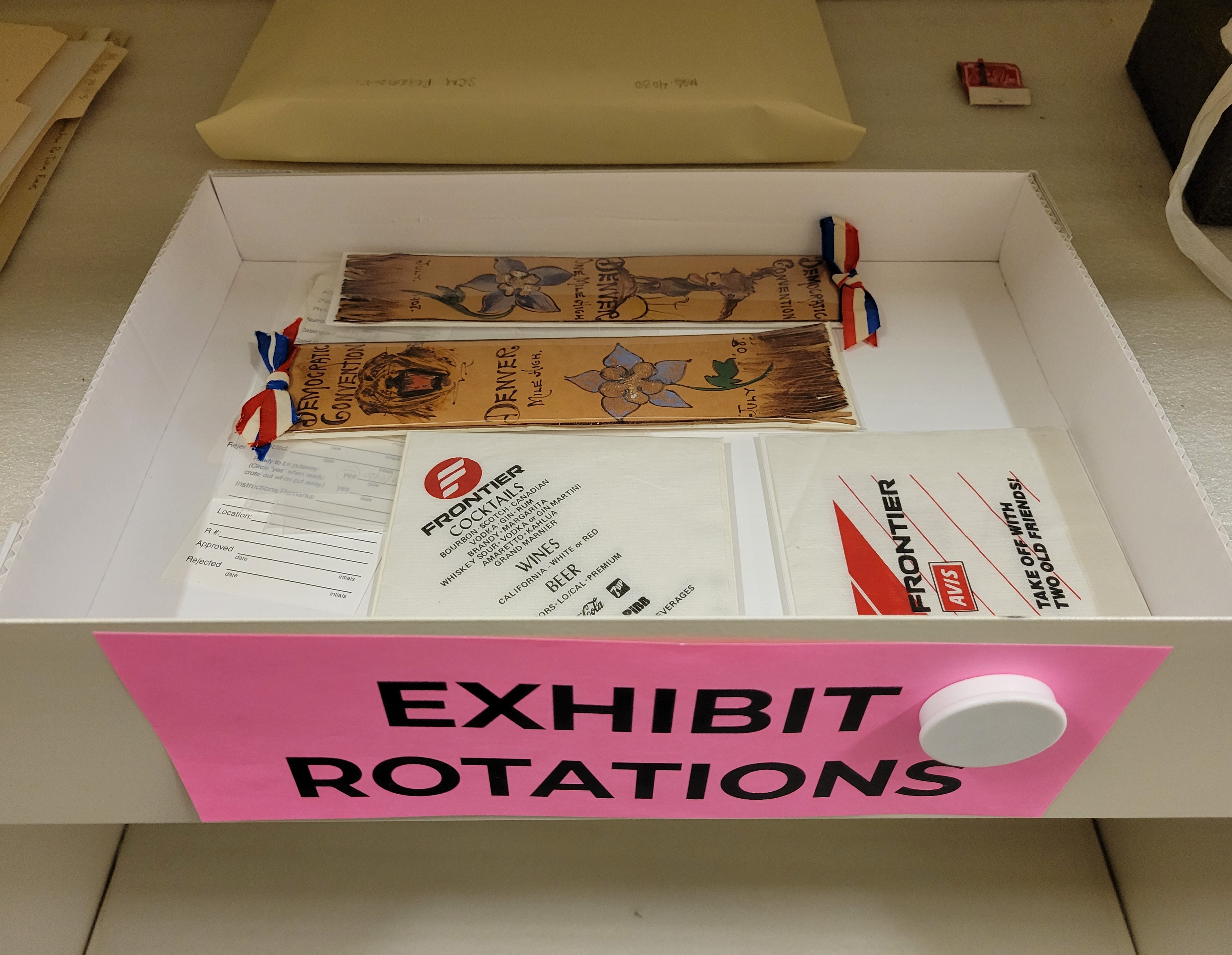 Photo of an open white box with a large pink sign on the front that reads "Exhibit Rotations." Inside of the box are two ribbons from the same Democratic Convention in Denver. Each brown ribbon has a red, white, and blue striped bow at the top. Beneath that is a handpainted animal and a columbine blossom at the bottom. Also in the box are two white paper cocktail napkins with the Frontier Airlines logo and black printed text. A museum registration slip lays next to them in the box. 