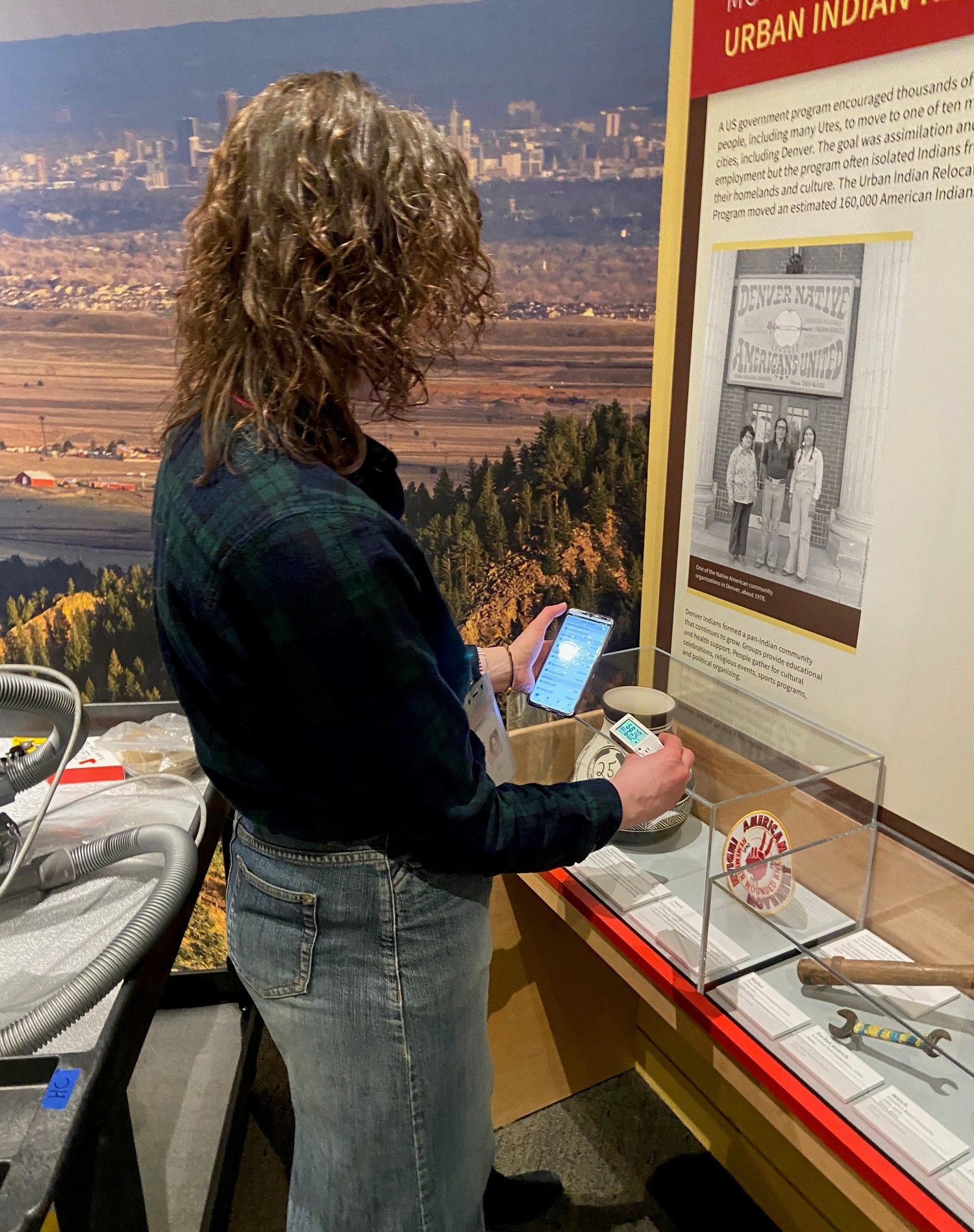 Photo of a museum staff member holding their smartphone in one hand and another snall electronic device in the other. They look to be comparing the data between the two devices in an effort to check the environmental readings of the exhibition environment.