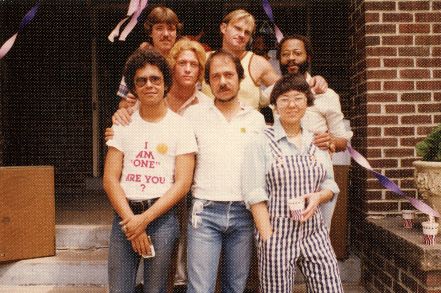 Photo of a group of people standing on the steps of a red brick building. There are streamers hanging from something behind them. The seven people are standing closely together, looking at the camera. One person in the front wears a white short-sleeved t-shirt that says "I AM 'ONE' - ARE YOU?" 