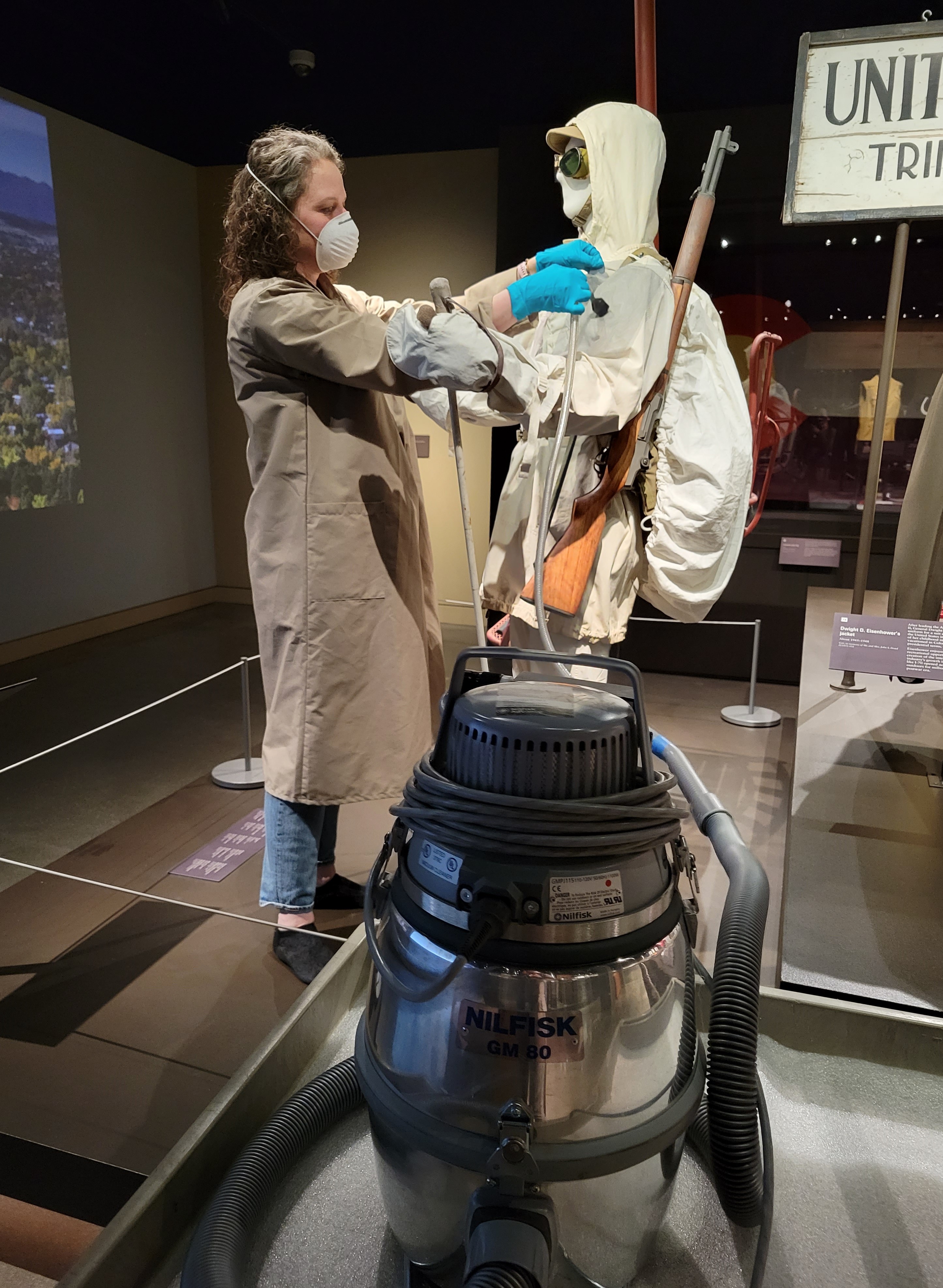 Photo of a museum staff member carefully vacuuming the textiles worn by a mannequin in one of the exhibits. The staff member is dressed in a long tan-colored coat with long sleeves, and they are wearing a white respirator mask over their nose and mouth. They use blue nitril gloves as they carefully handle the textile artifacts. In the foreground, the stainless steel vacuum is visible, its long hose reaching across to the mannequin that is dressed in a white 10th Mountain Division ski suit.