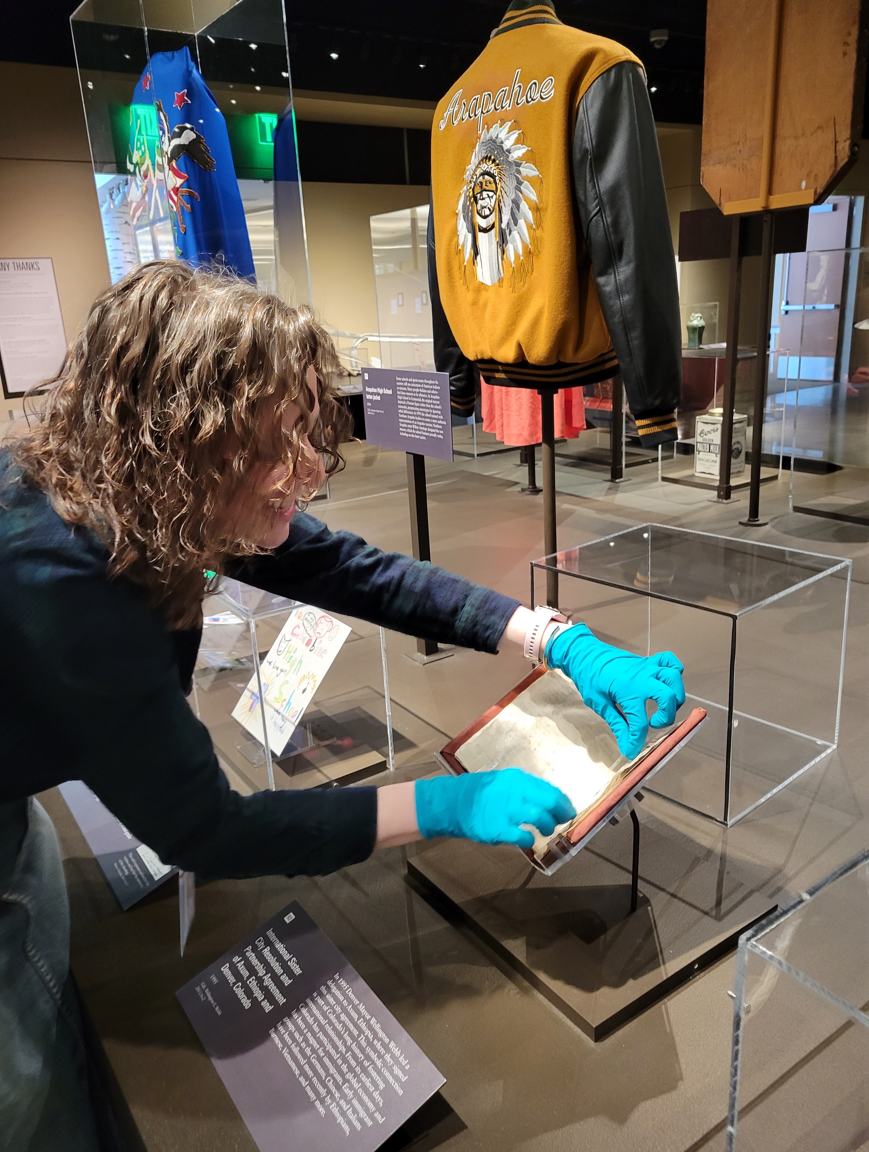 Photo of a member of museum staff, carefully caring for the pages of a document that is on display in the Zoom In exhibition. The staff member wears blue nitril gloves to handle the pages of the document. 