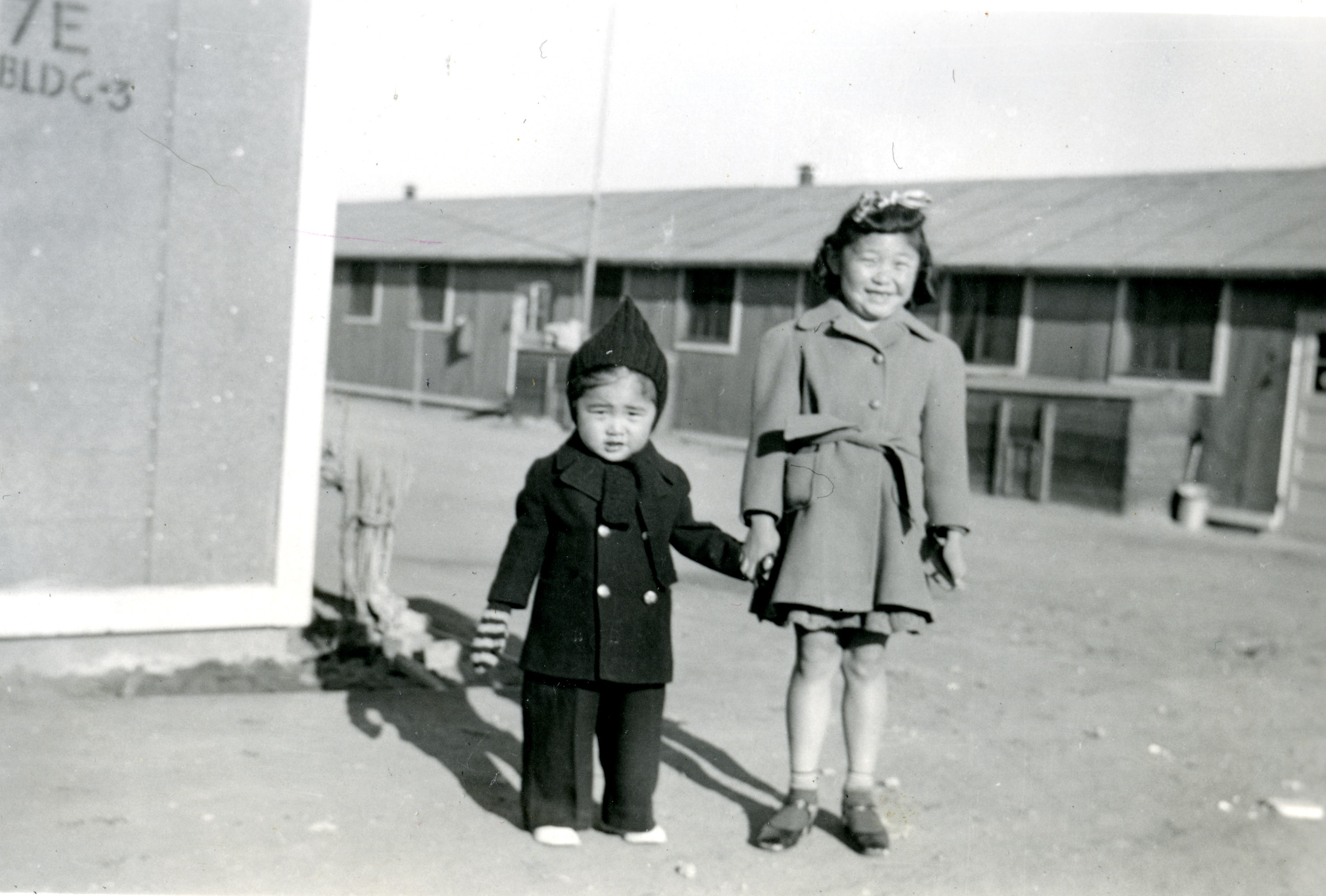 Two children, dressed for cold weather, pose before a barracks at the Amache incarceration camp.