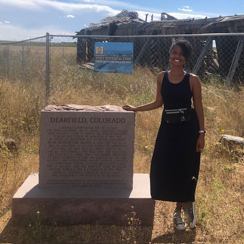 Photo of SHF Staff Member, Sophia Felder, at the Dearfield site in Colorado. A plaque dedicated to Dearfield is to Sophia's right, and a historic structure from Dearfield is in the background.