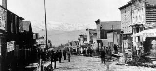 Black and white image of State Street in Leadville, Colorado. Many buildings line the debris-filled street, with Rocky Mountains in the background. 