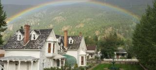 A white house on the left with a well manicured green lawn on the right and a green fountain in the center of the lawn. A rainbow crosses the entire picture above the house intersecting the mountains in the background.