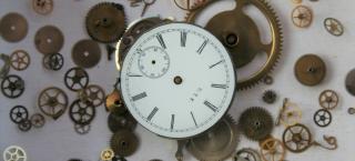 Photo of a white clock face with thin black roman numerals. The clock is in pieces, and all of the small cogs that make the clock function are scattered around in the area around the clock face.  The hands on the clock face are missing.