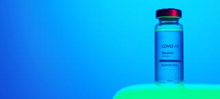 Photo of one vial of Covid-19 Vaccine. The Vial has a red metal top and a label that says "COVID-19 Vaccine, 2019 n-CoV, Injection Only." The vial sits atop a green table, and the background is a luminescent blue.