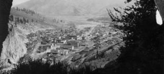 Photo of the town of Creede in the year 1918. The image is taken from atop a hill overlooking the town, so trees and brush frame the view of the town in the distance. Two main streets through town are evident, and low buildings of small to mid-sized are arranged through the town. No vehicles appear to be seen, and the town is too far away to see people walking around..