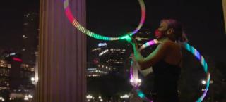 Photo of a woman in the middle of a hula hoop performance. She is using two hoops of different sizes, which are illuminated by multicolored LED lights. She is wearing a black sleeveless leotard and she is wearing a mask as a coronavirus precaution.