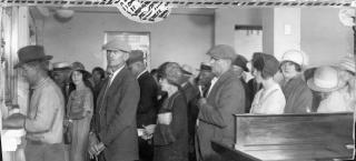 A photo of a queue of customers at Broadway National Bank in Denver. A man is conducting business at the teller window to the left of the image, while several people are waiting quietly in line behind him, many holding a handbag or documents.
