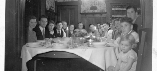 Photo of a large family seated closely together around a holiday meal table. On the far side of the table there is a turkey, and two candles decorate the table. Relatives of all ages smile for the group photograph before beginning their meal.